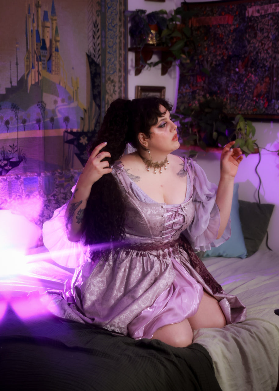 Woman wearing lavender floral satin corset dress with puffed sleeves sitting on bed in softly lit room