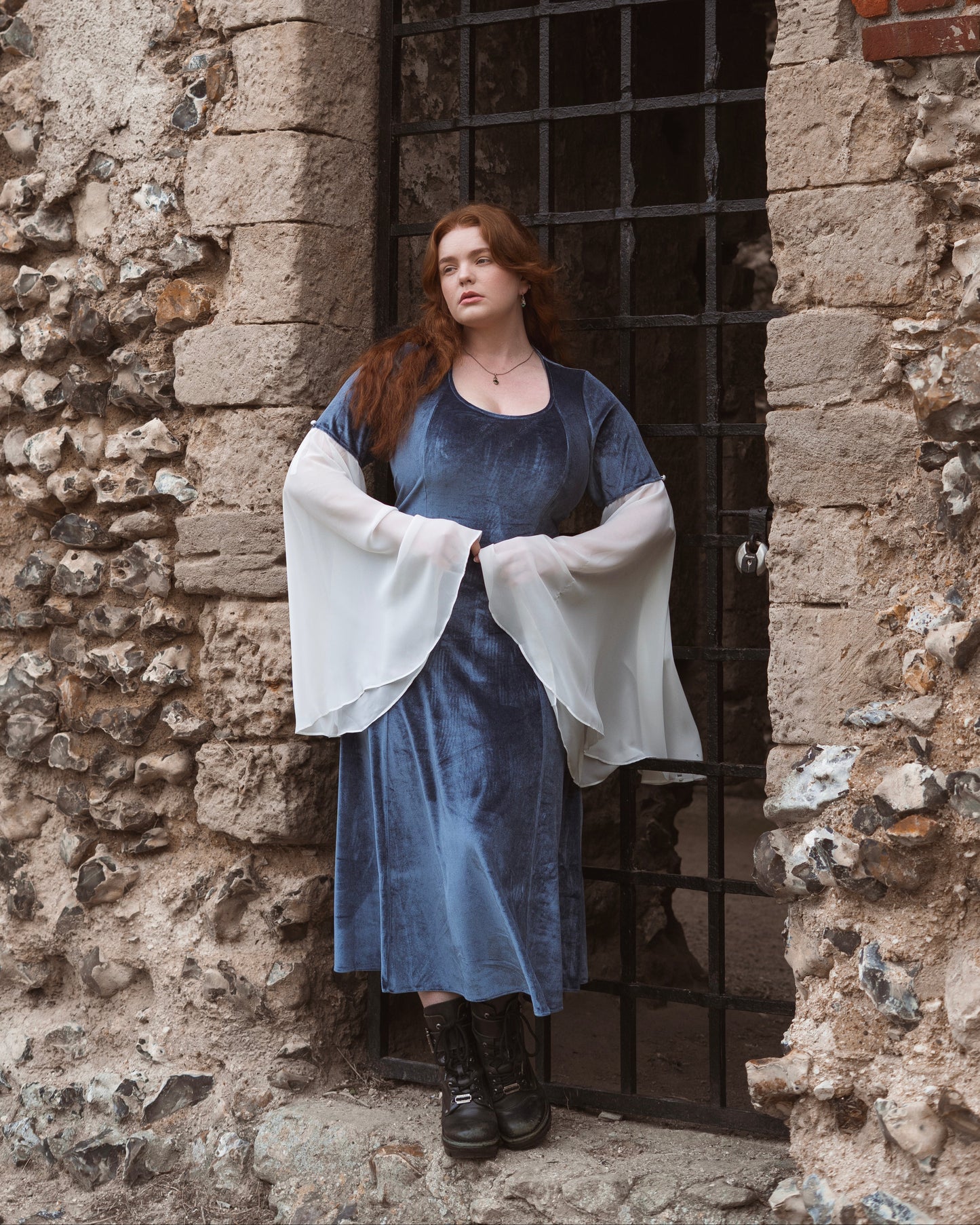 Woman wearing Elven Blue Velvet Bell Sleeve Dress with sheer white flowing sleeves standing by stone archway and metal gate
