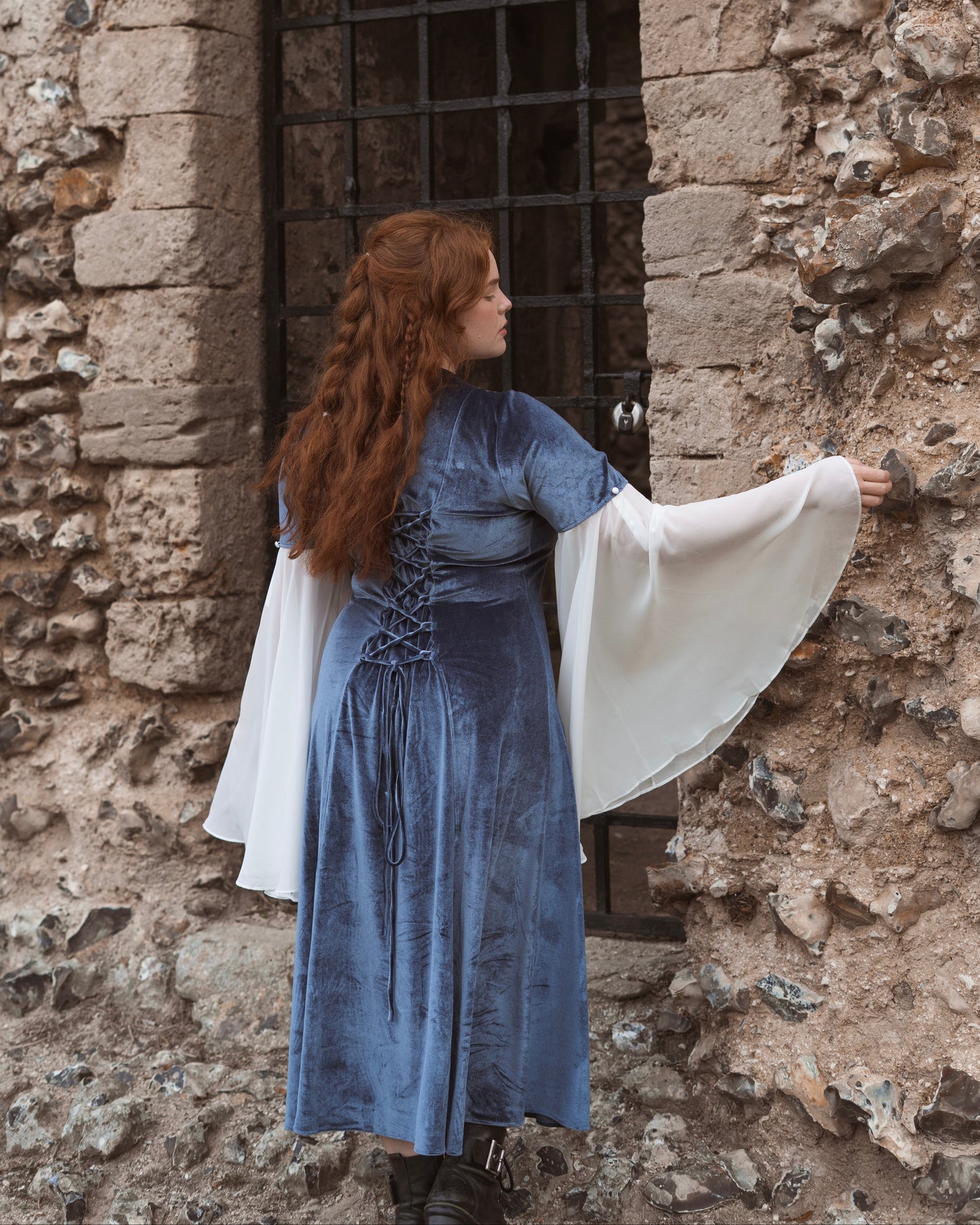Woman wearing Elven Blue Velvet Bell Sleeve Dress with adjustable back lacing standing by rustic stone wall and iron gate