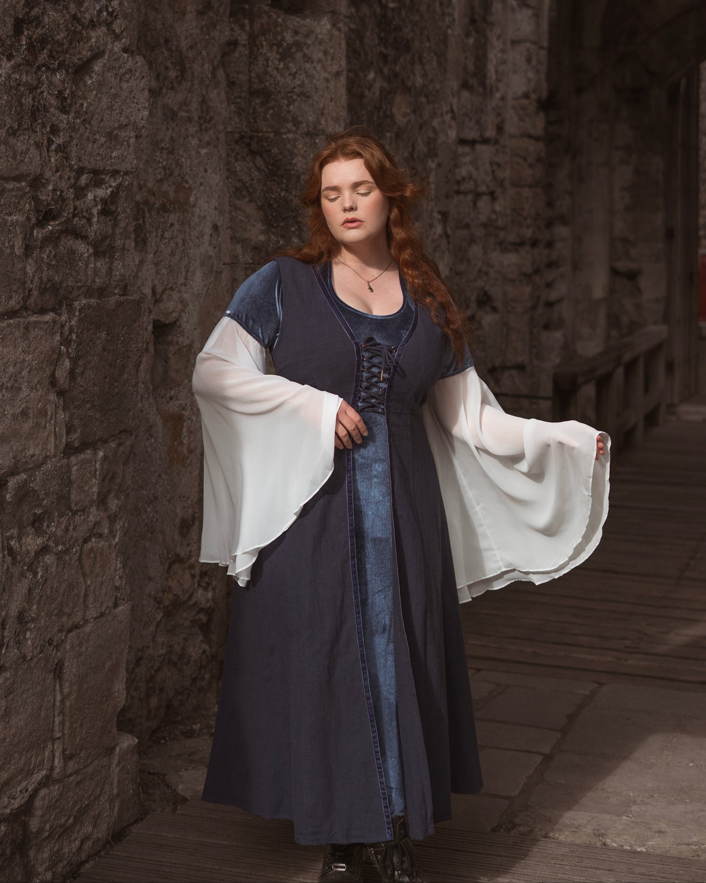 Medieval-inspired navy blue kirtle dress with empire waist and flowing white sleeves modeled in stone corridor.