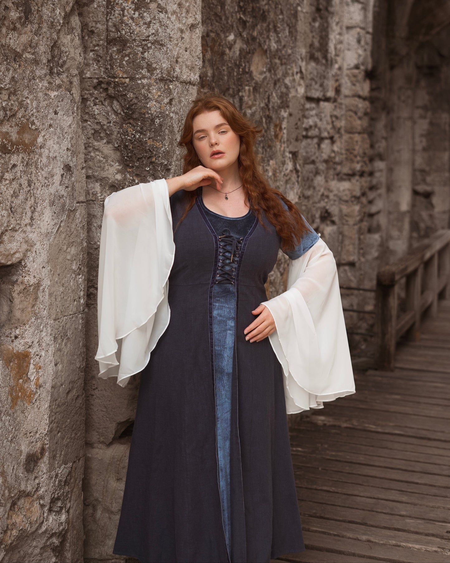 Woman wearing a medieval style dress with bell chiffon sleeves and adjustable back lacing standing by stone wall