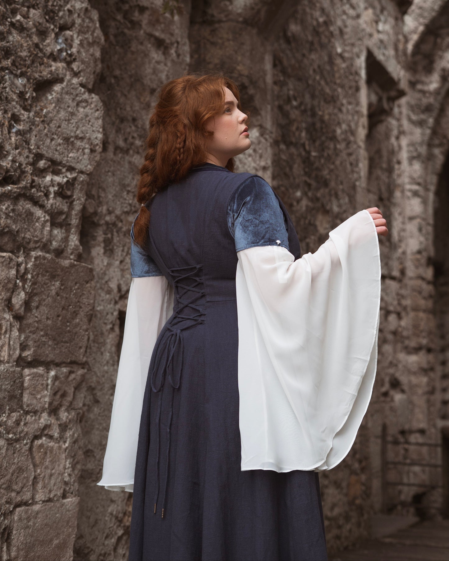 Woman wearing a navy blue Medieval-style kirtle dress with laced back and flowing white sleeves standing by stone walls