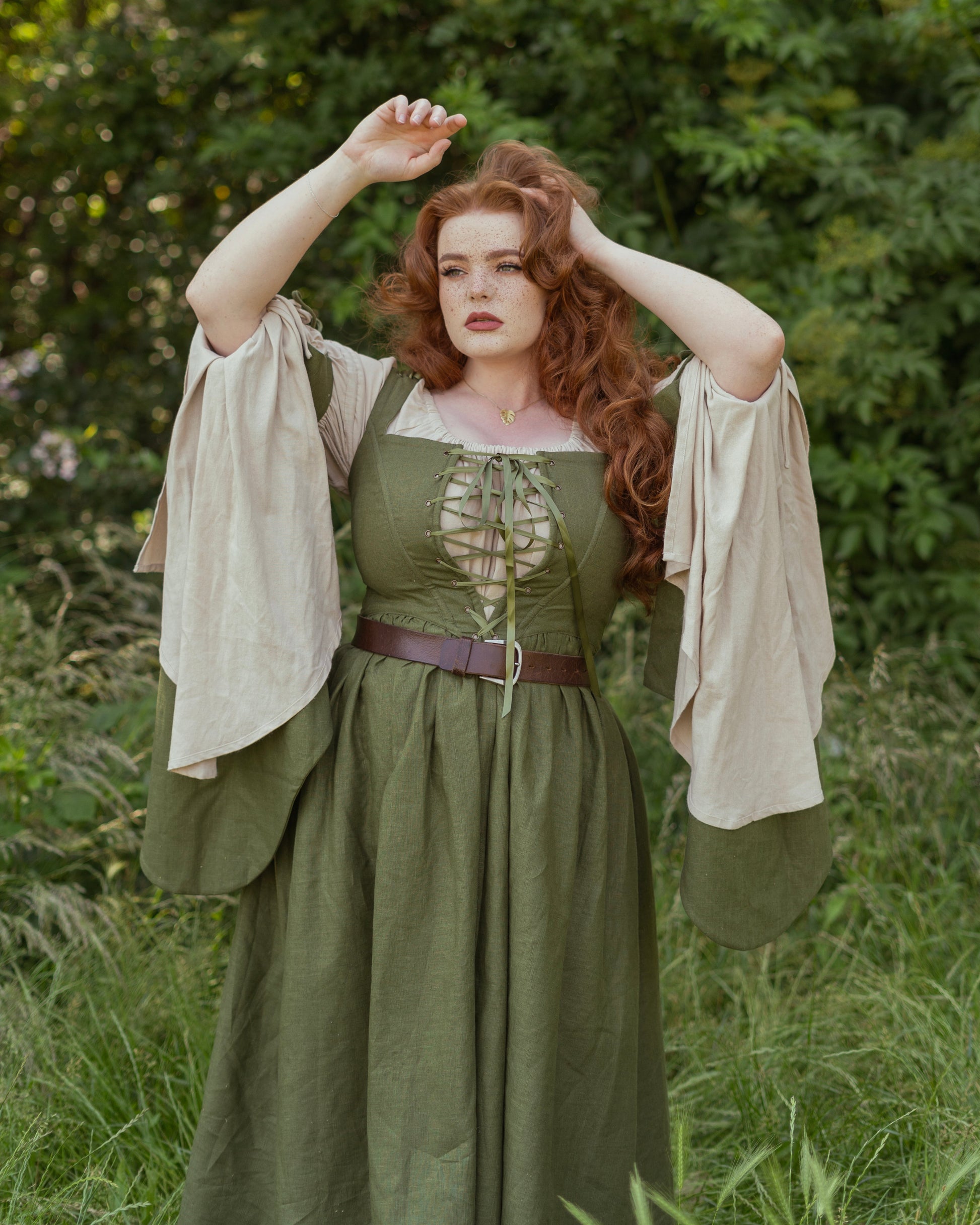 Woman wearing ivy green linen corset dress with flowing bell sleeves in a natural woodland setting