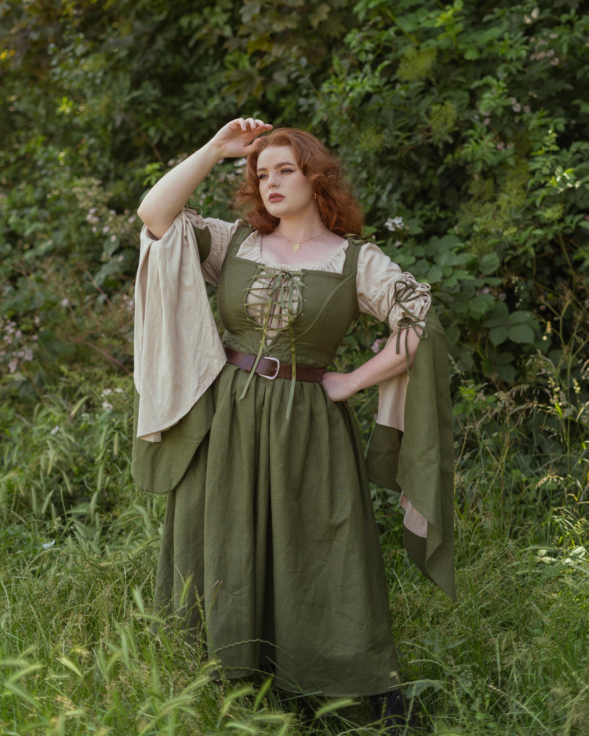 Woman wearing an ivy green linen corset dress with flowing bell sleeves in a lush forest setting