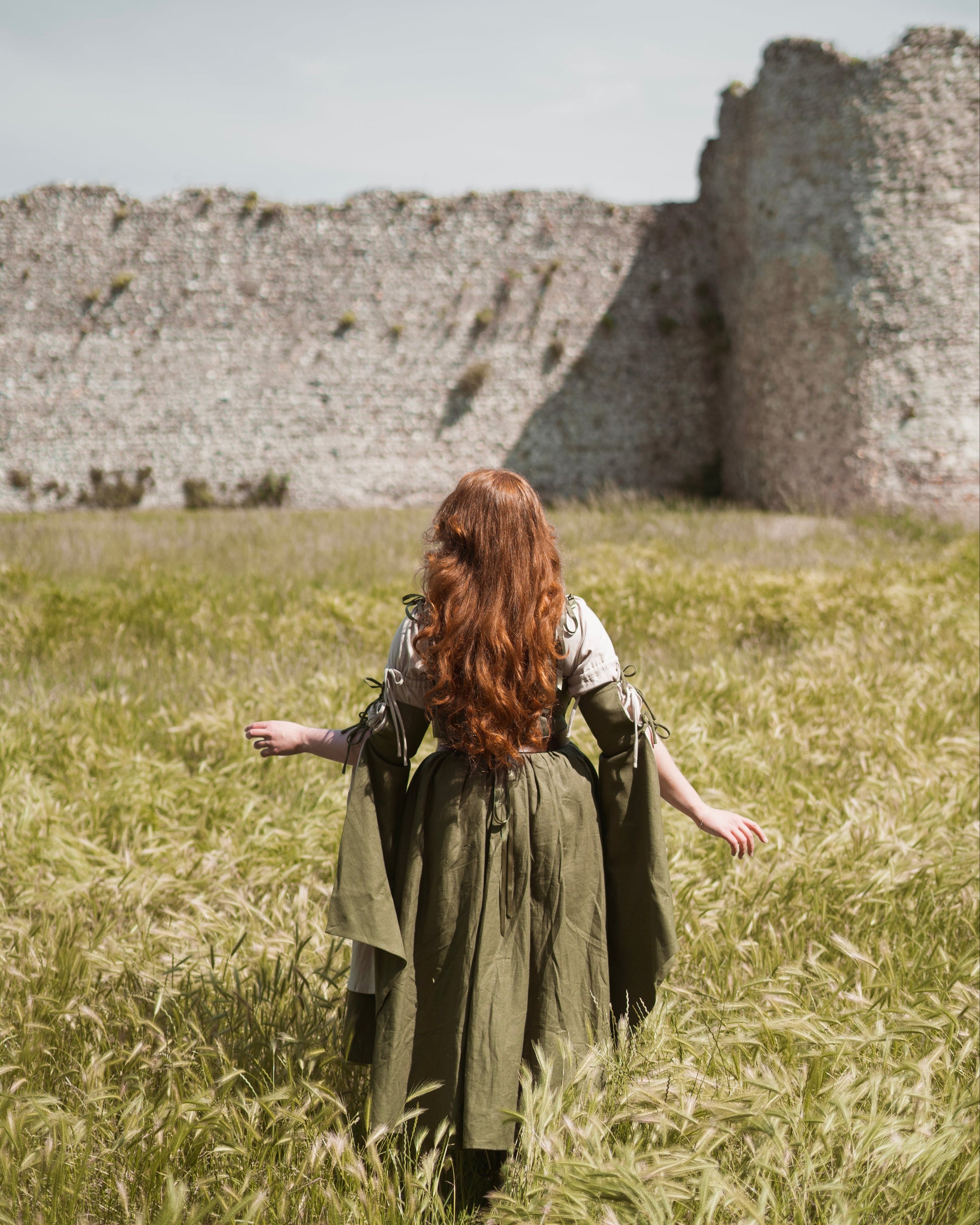 Woman with long red hair wearing an ivy green linen corset dress with flowing sleeves in a grassy field near ruins