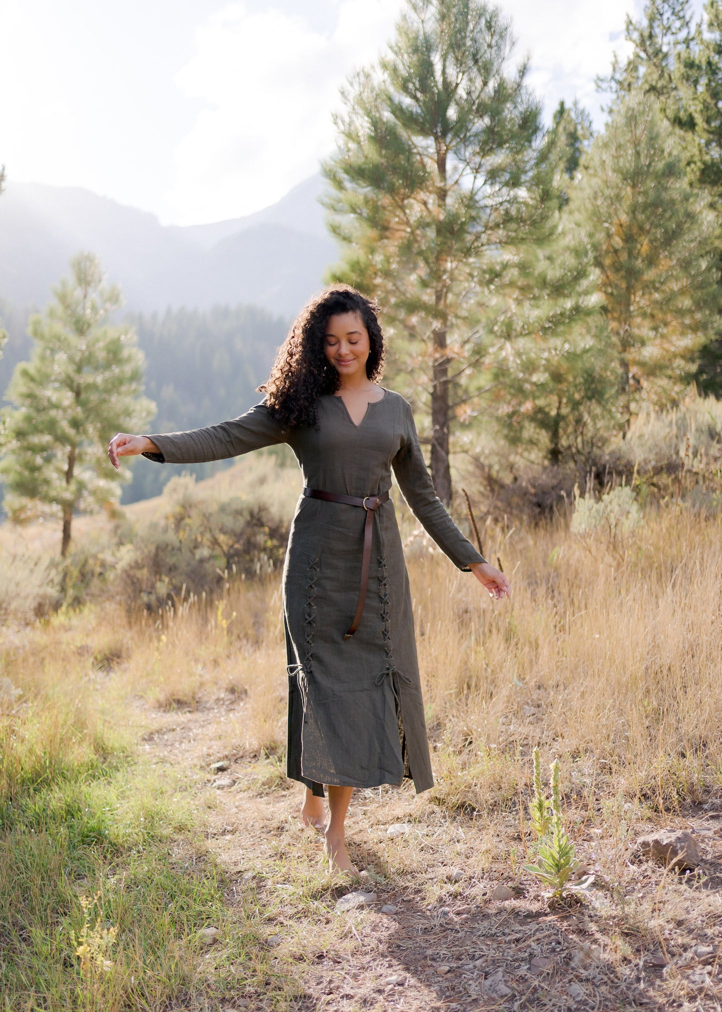 Olive Green Viking Dress shown outdoors in mountain setting with laced front detail and long sleeves on nature trail with pine trees
