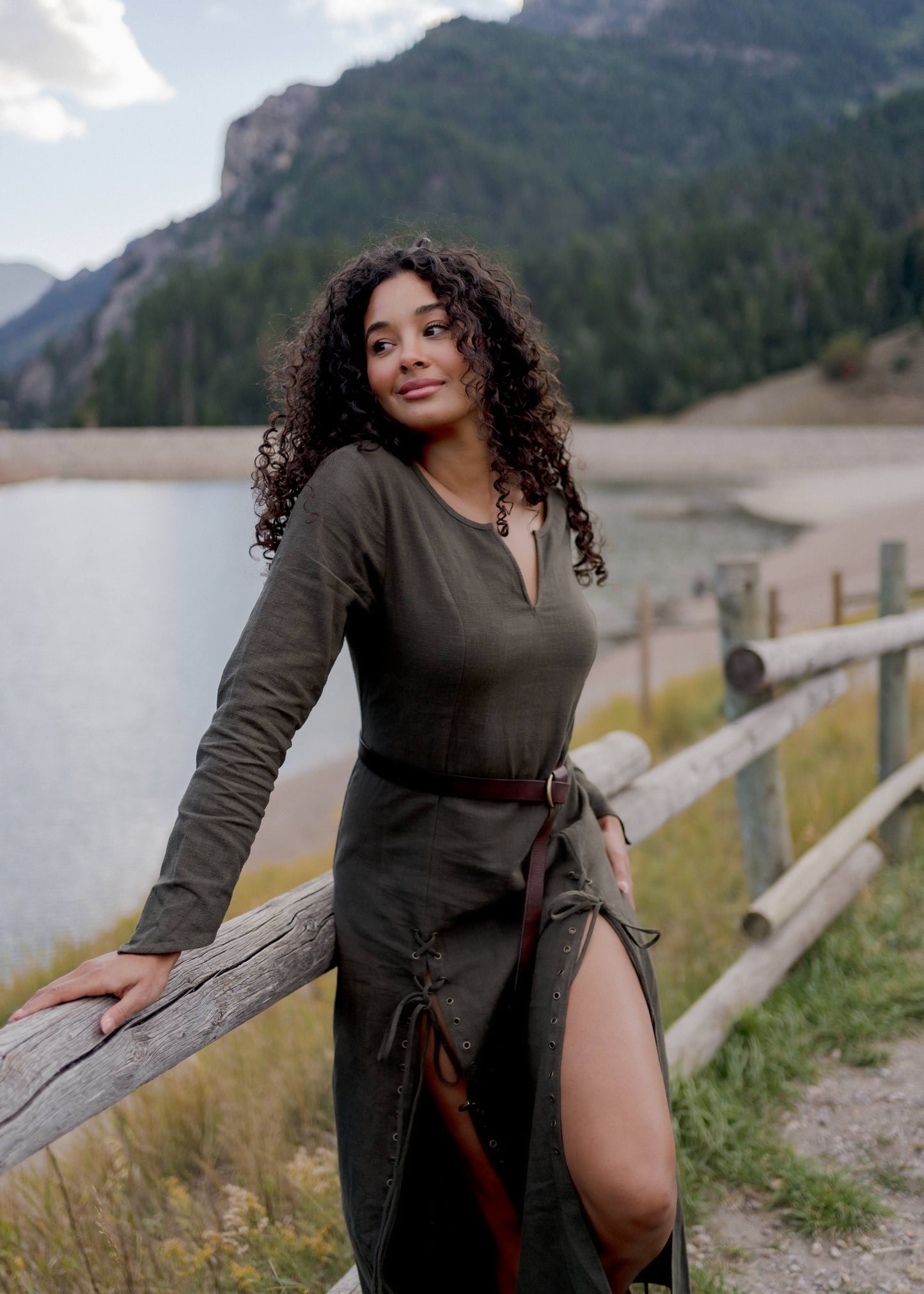 Olive Green Viking Dress shown outdoors by lake with mountains, featuring fitted silhouette, side slit, and leather belt detail