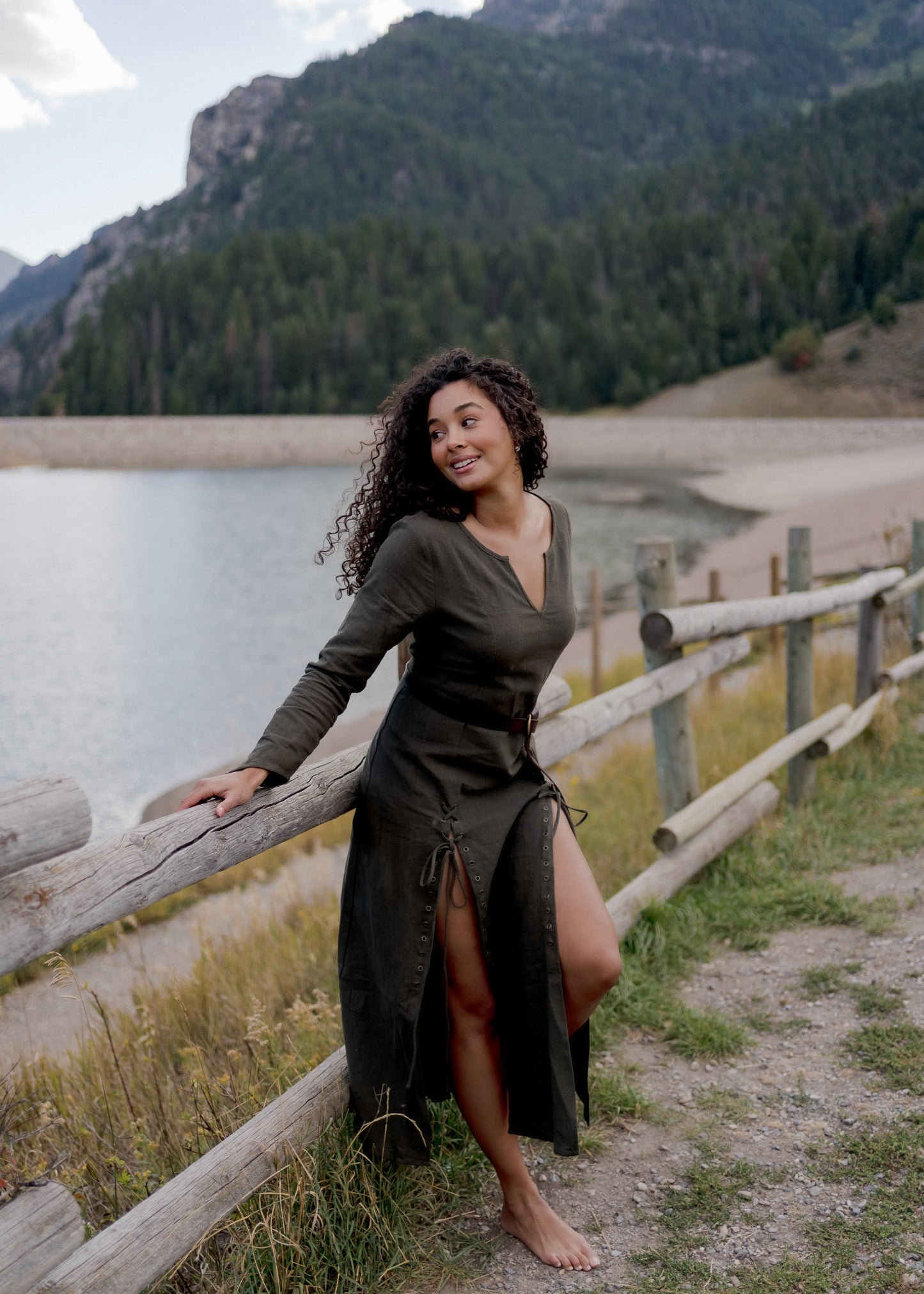 Olive Green Viking Dress with lace-up details shown outdoors by mountain lake with wooden fence, displaying front slit feature