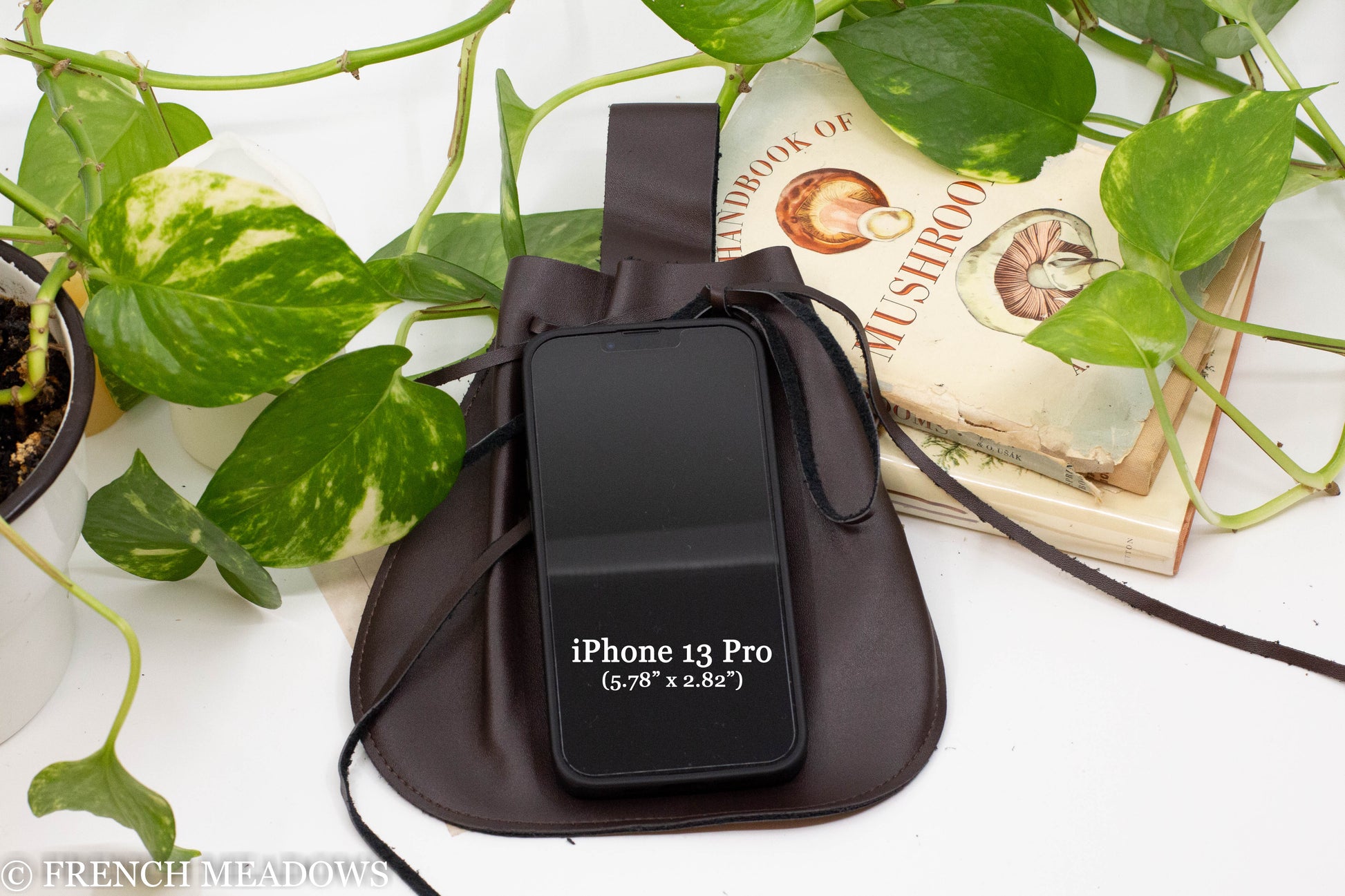 Brown vegan leather drawstring pouch next to books and an iPhone 13 Pro for size reference, set among green plants