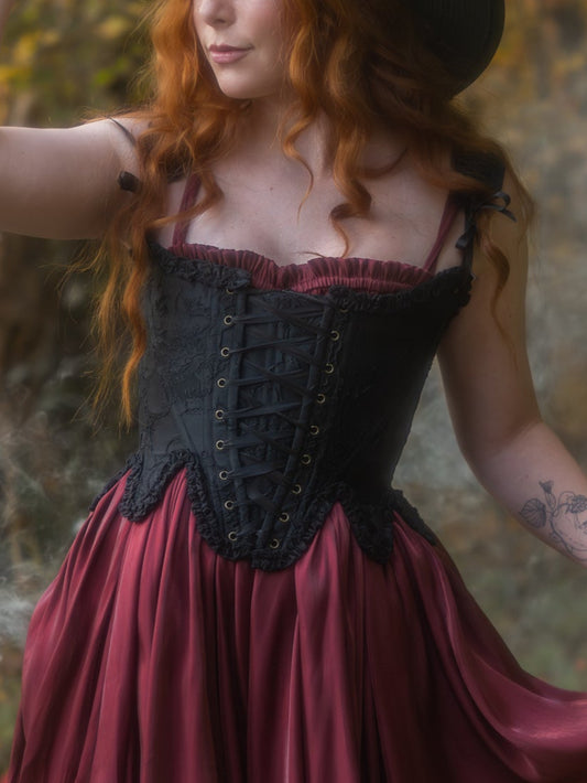 Woman wearing black floral jacquard 18th century stays with ruffled neckline and maroon skirt outdoors