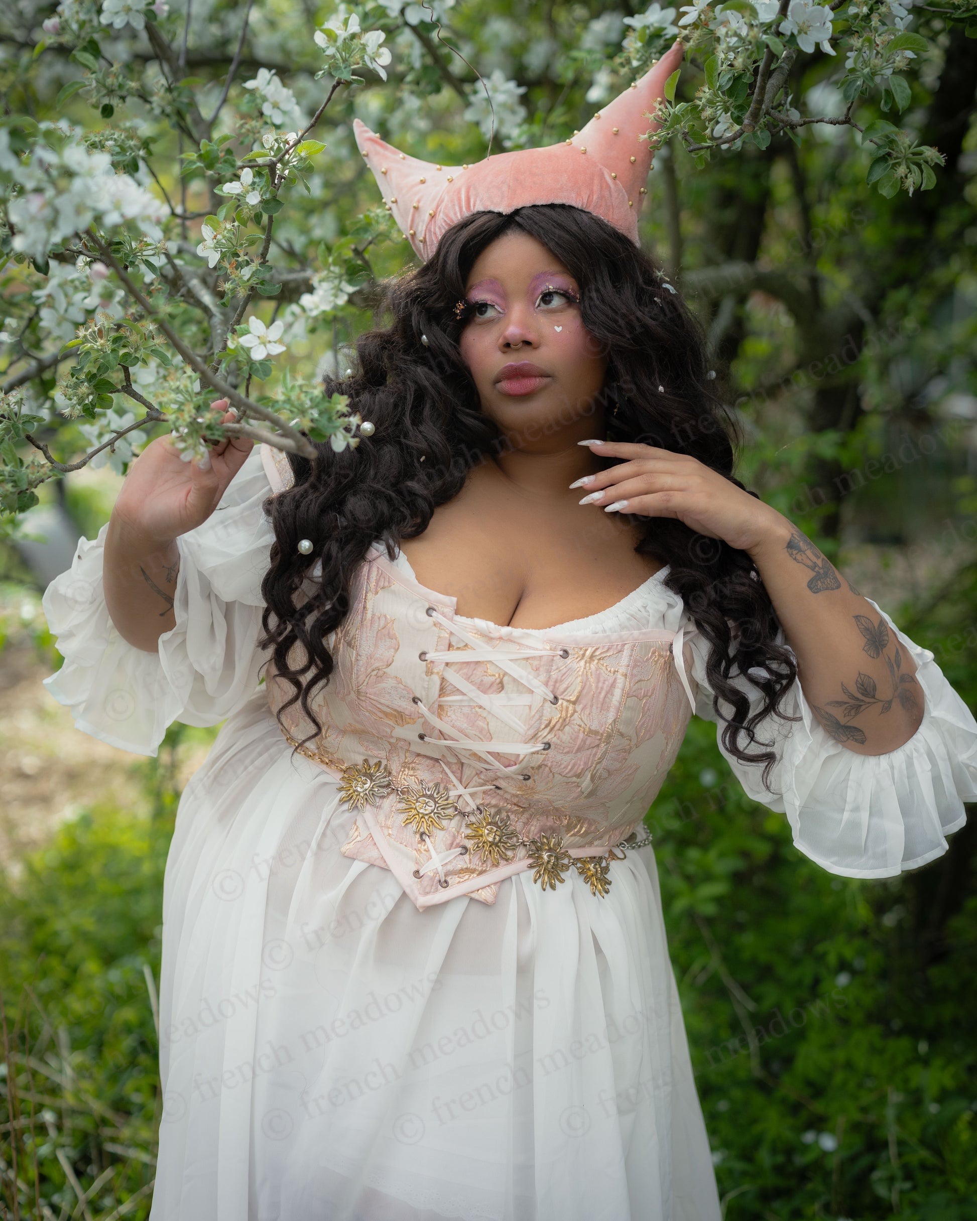 Curvy woman wearing a lace-up Renaissance corset and flowing white dress posing by blossoming tree branches.