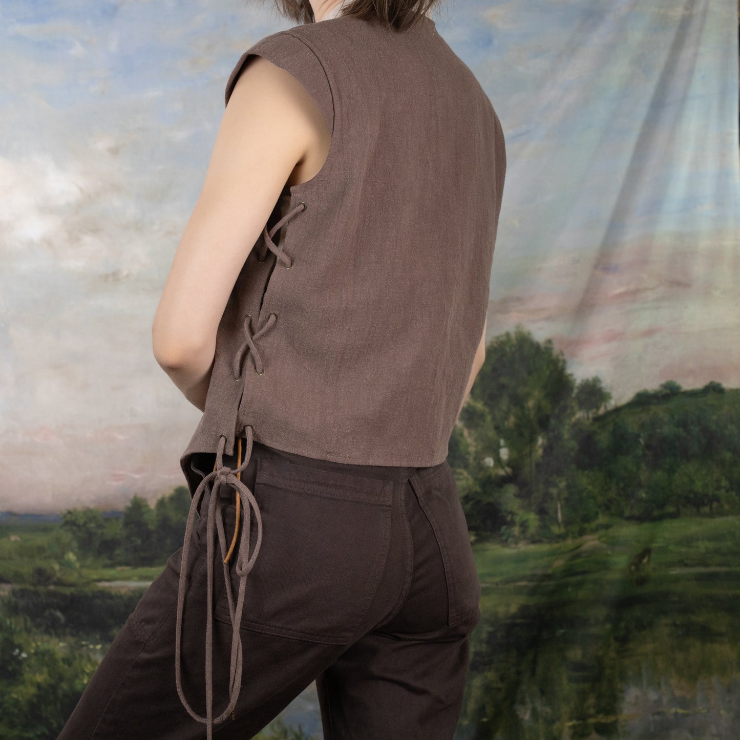 Brown Bard Vest with side lacing detail and adjustable ties shown from back view against pastoral landscape backdrop