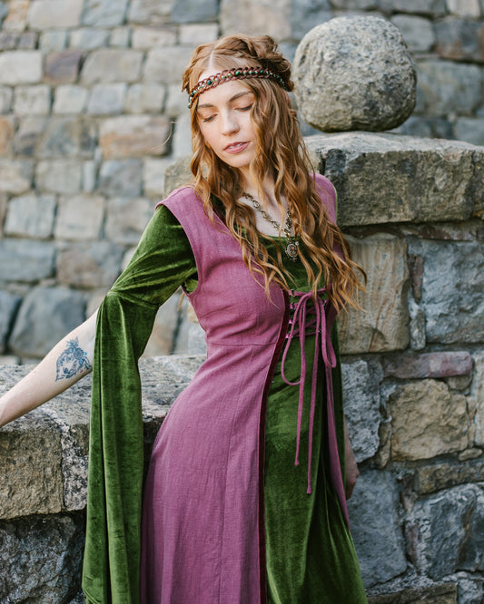 woman wearing a green velvet medieval dress with long bell sleeves and a purple corset-style overlay outdoors by stone wall