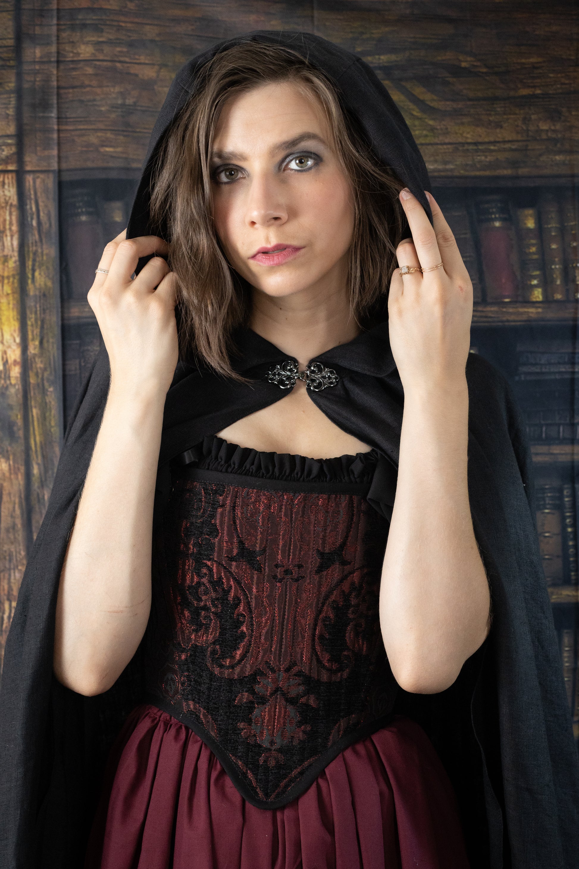 Black linen renaissance cloak worn over burgundy corset dress against vintage library backdrop, featuring ornate clasp