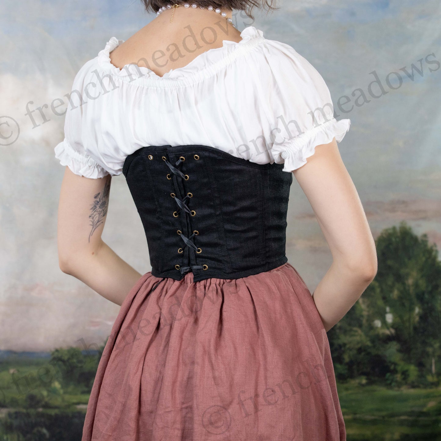 Black cotton corset top with lace-up back detail shown with white peasant blouse and dusty rose skirt against pastoral backdrop