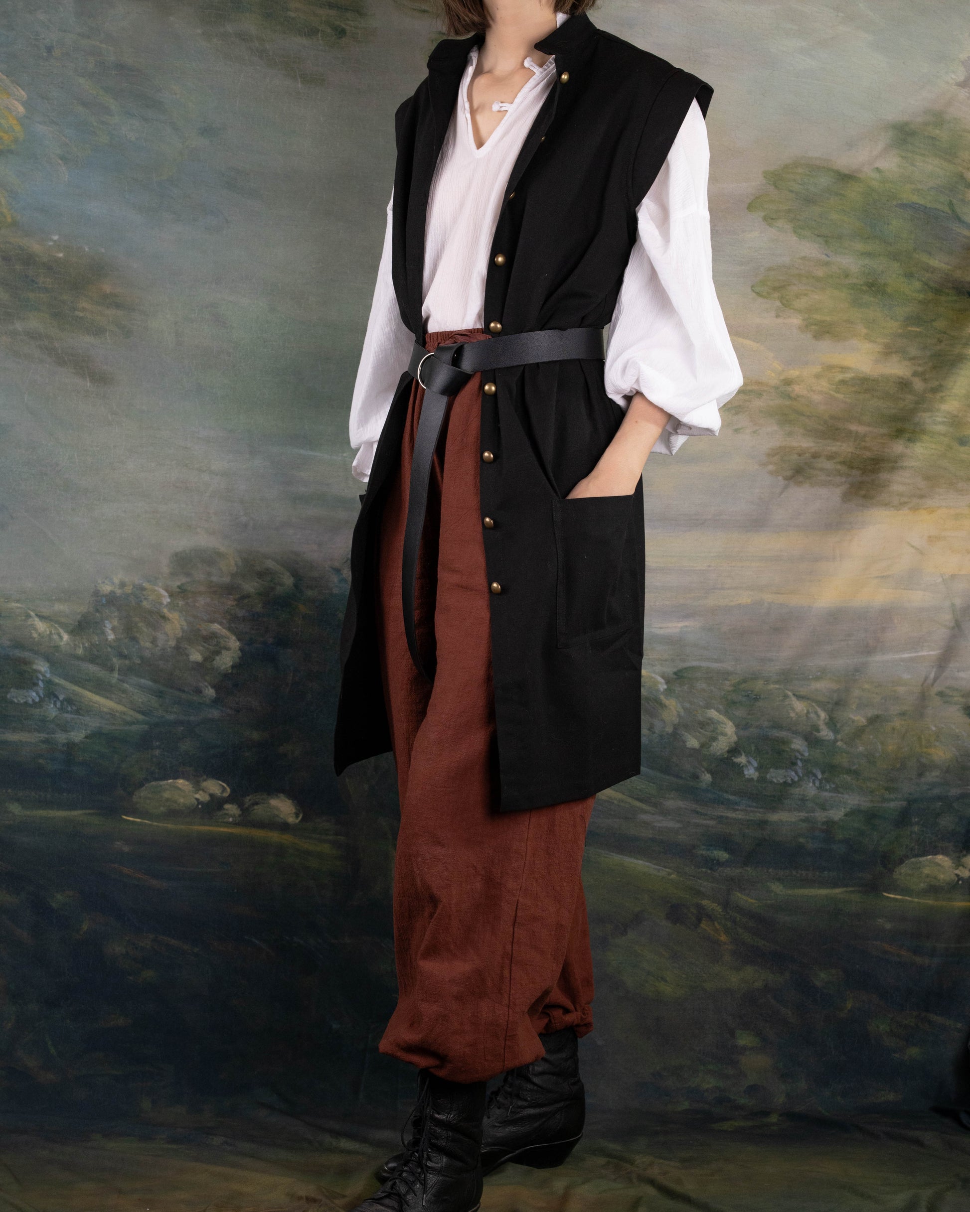 Model wearing Black Pirate Vest over white shirt and brown pants with black belt and boots in renaissance style setting.