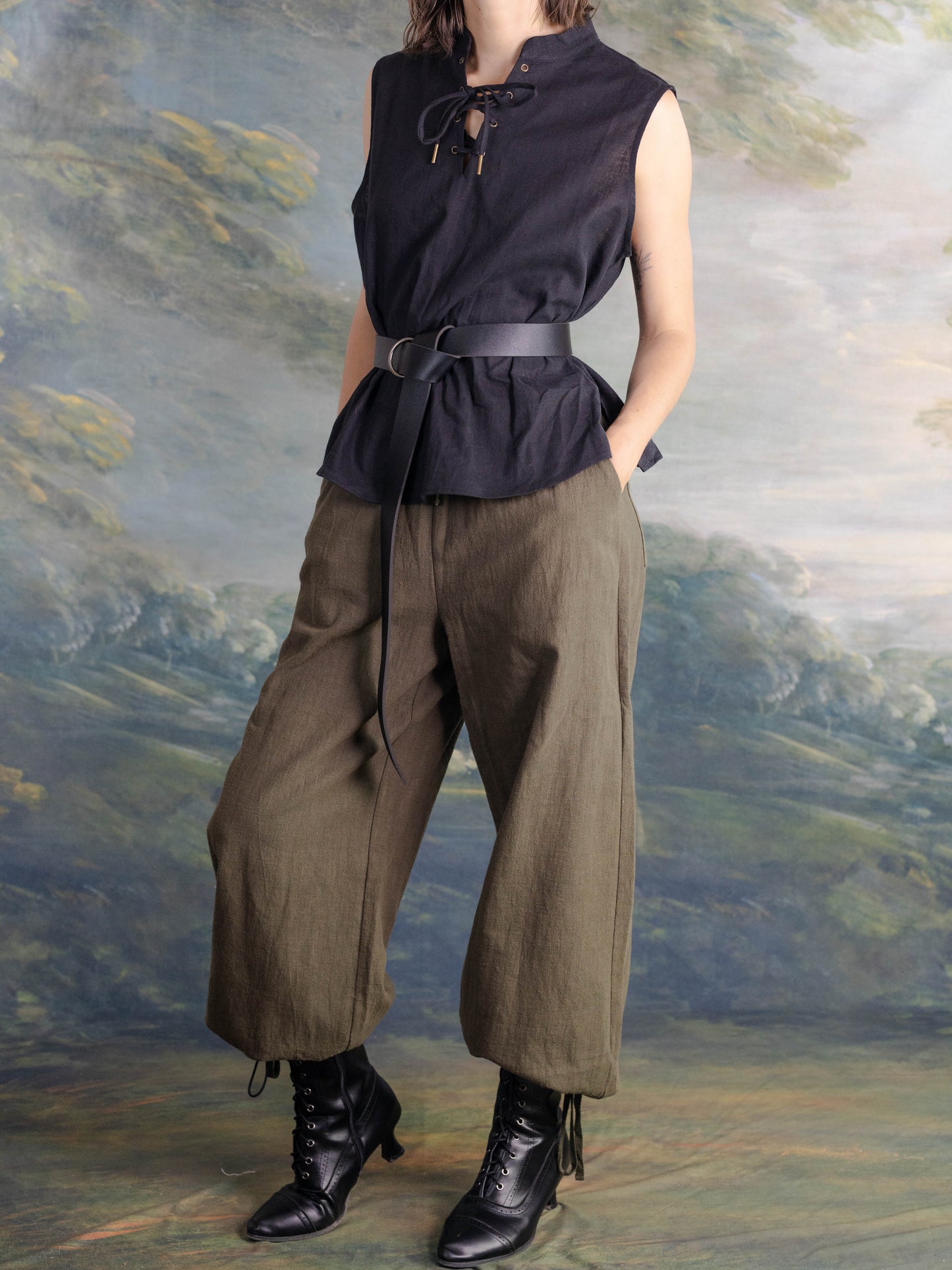 woman wearing sleeveless black pirate tank with lace-up neckline and olive wide-leg pants against painted backdrop