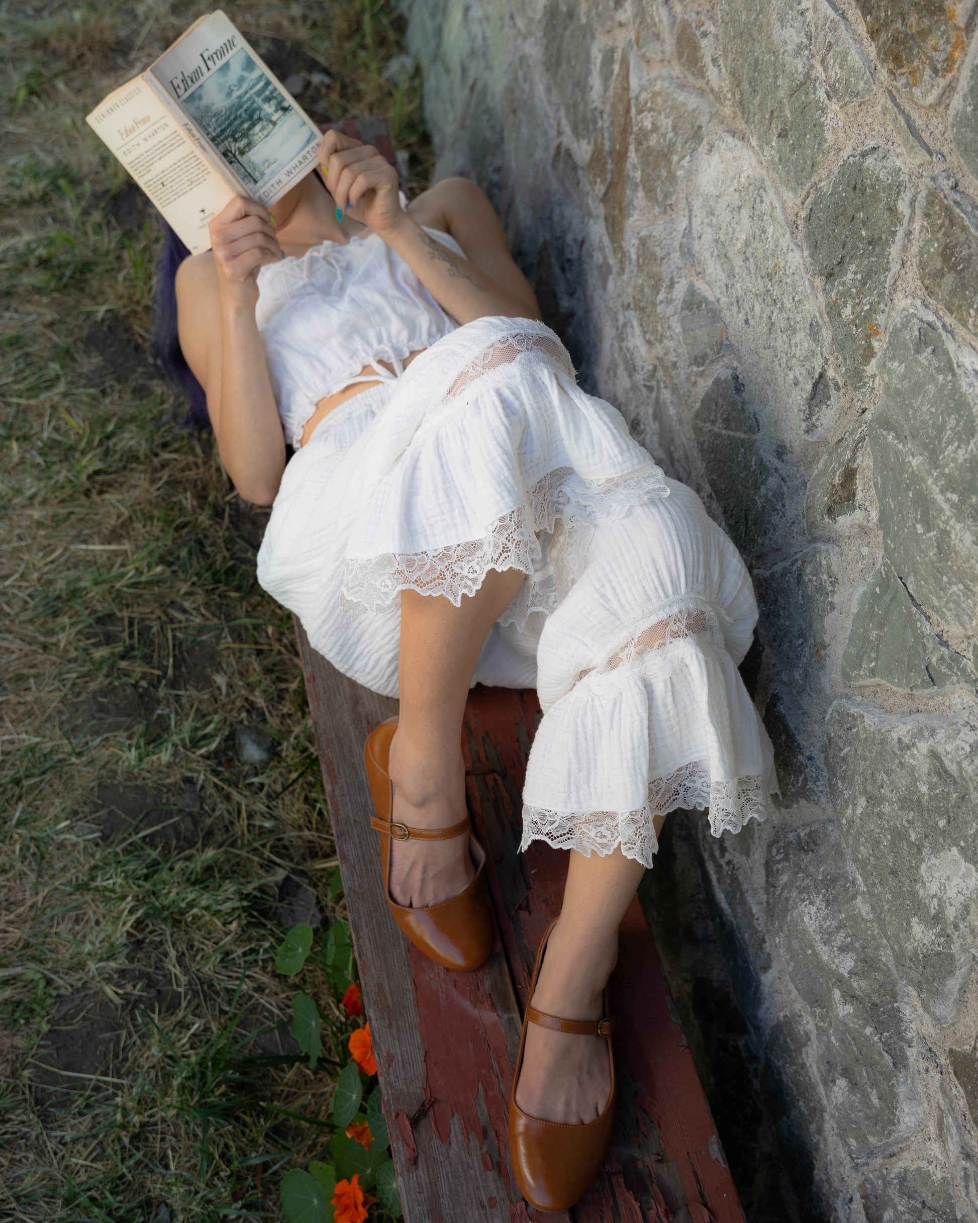 Woman wearing white cotton bloomer pantaloons with lace trim, lying on bench and reading a book by stone wall outdoors