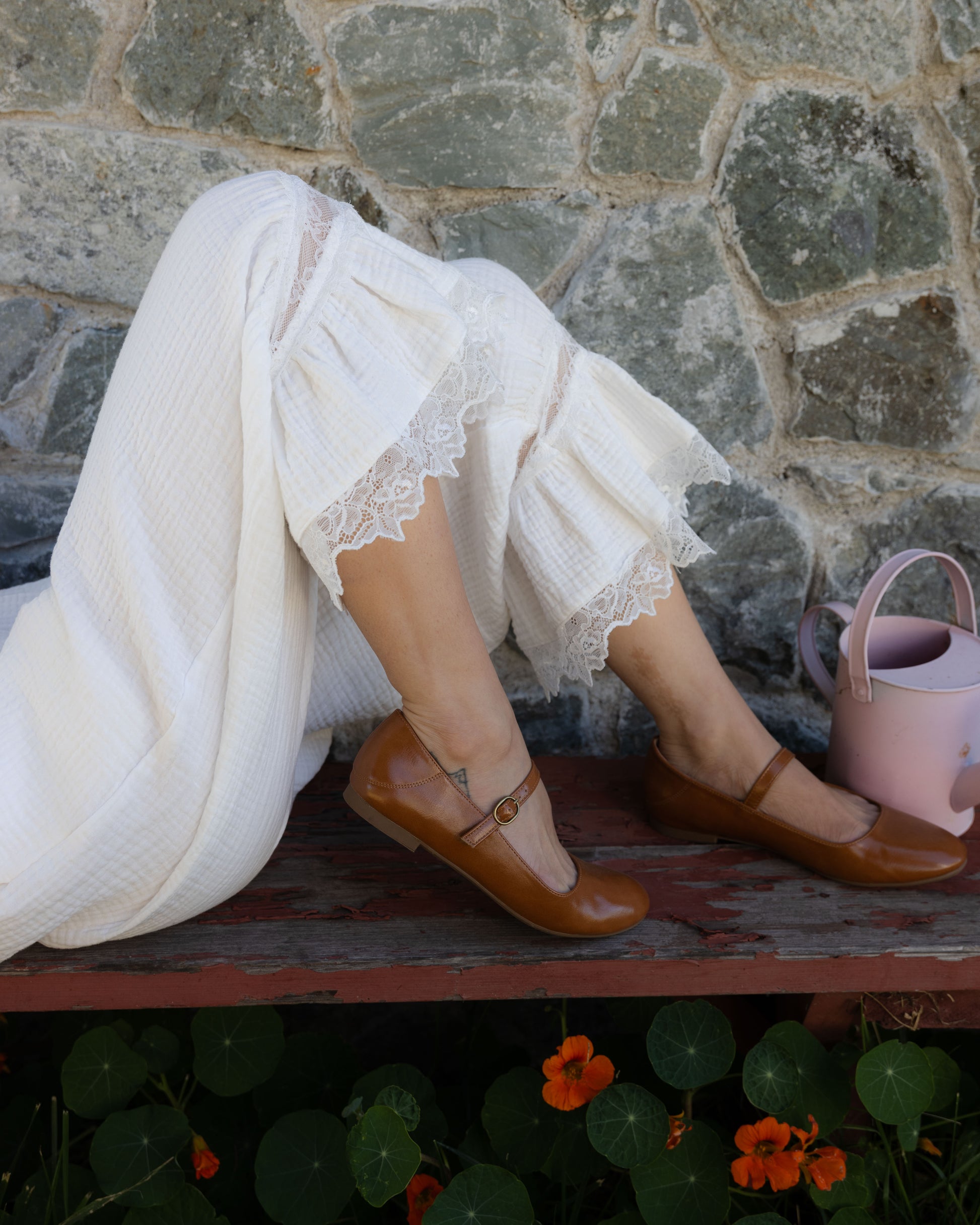 Woman wearing soft white bloomer pantaloons with lace trim, sitting on a bench outdoors next to a pink watering can