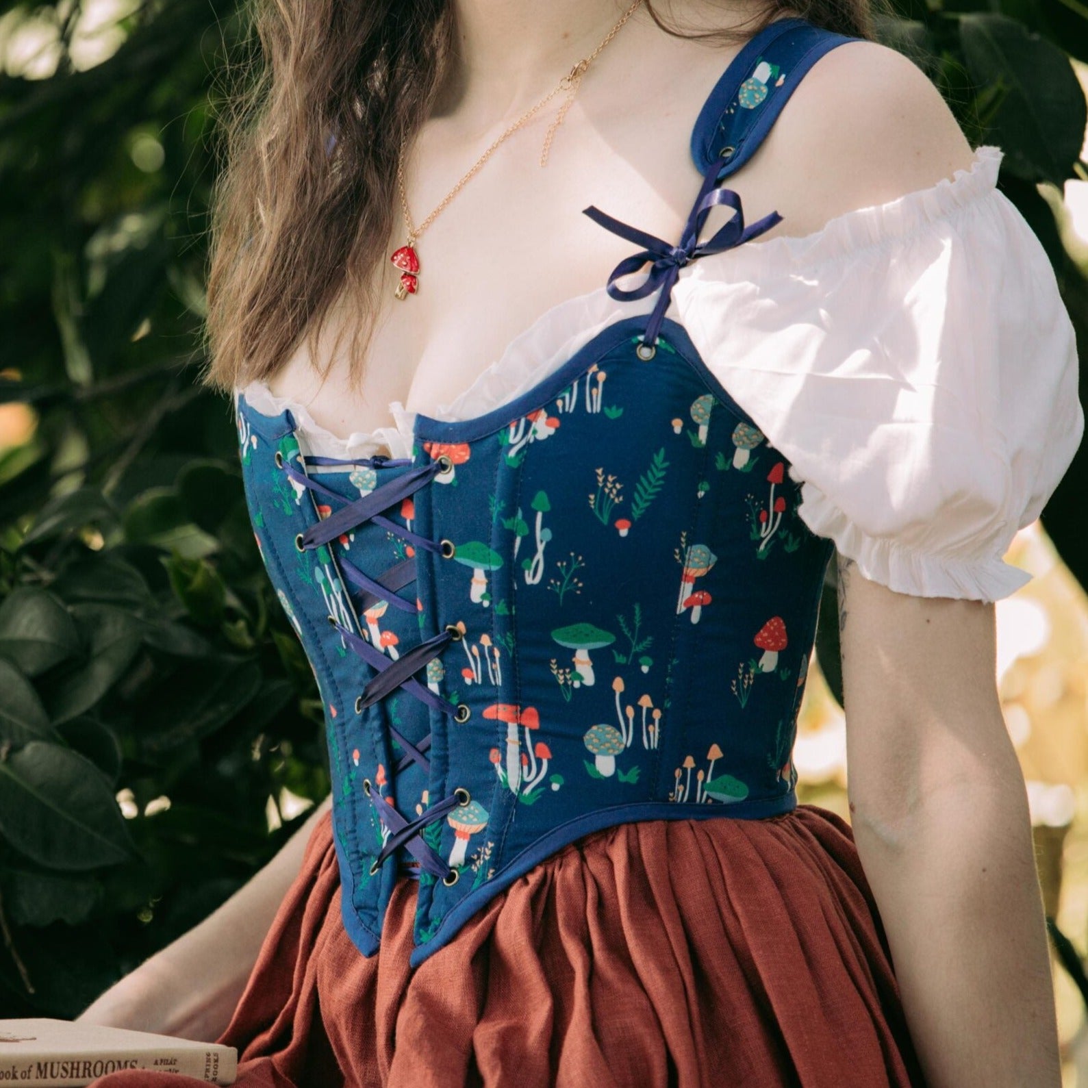 Blue Mushroom Renaissance Bodice with whimsical forest print, lace-up front and puff sleeves, worn with brown skirt in garden setting