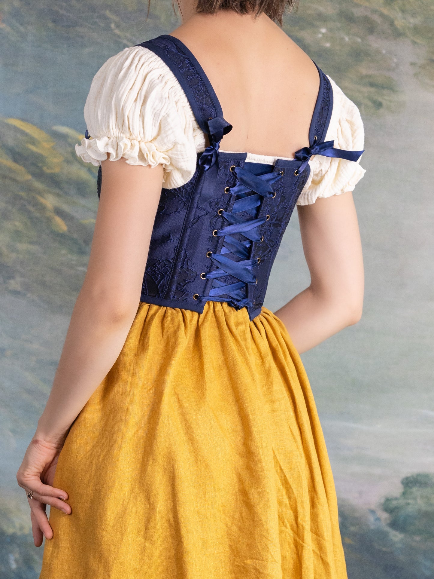 Back view of a woman wearing a dark blue floral jacquard corset laced up over a white blouse and yellow skirt