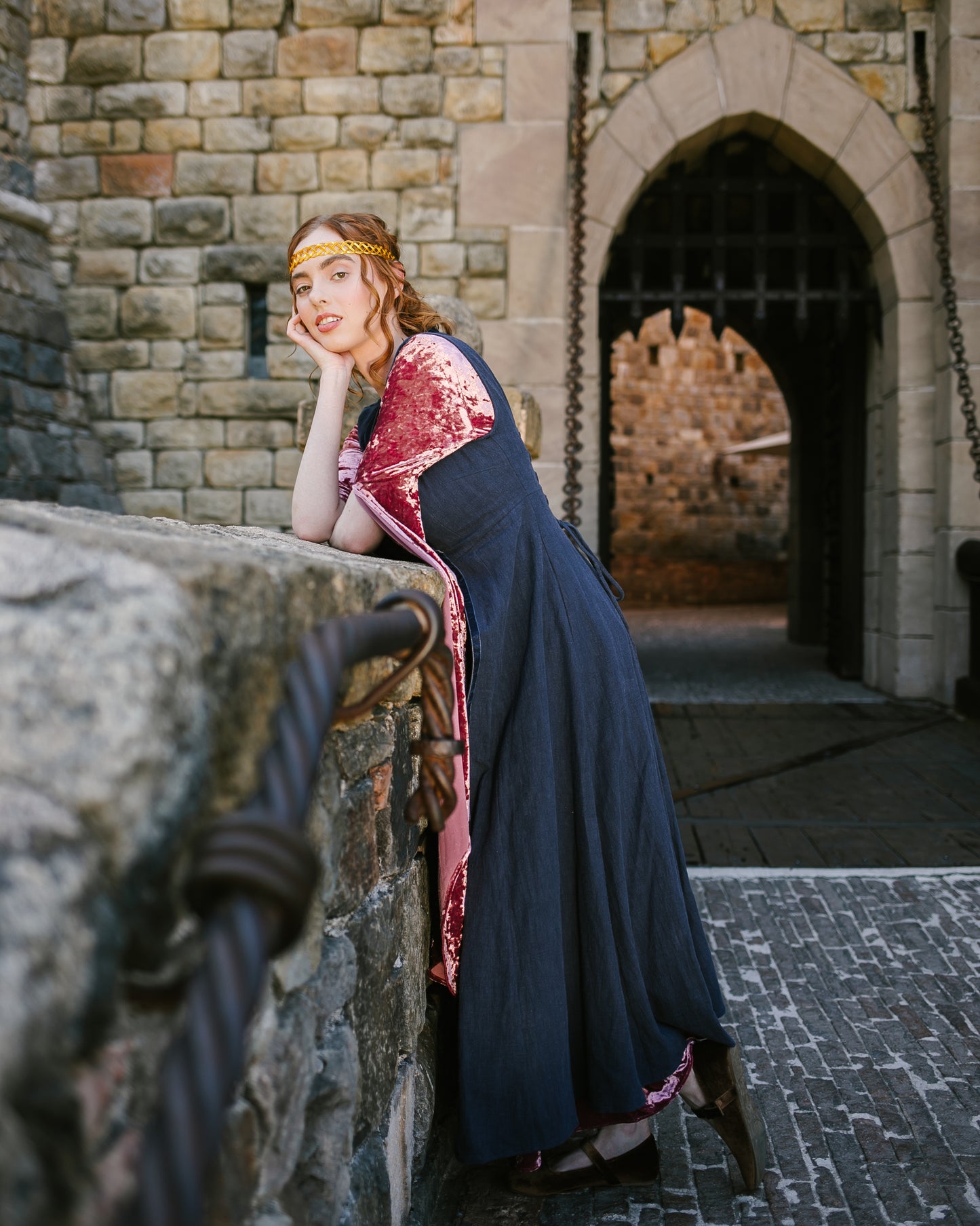 Woman wearing a navy blue Medieval kirtle dress with velvet sleeves leaning on a stone wall near a castle entrance