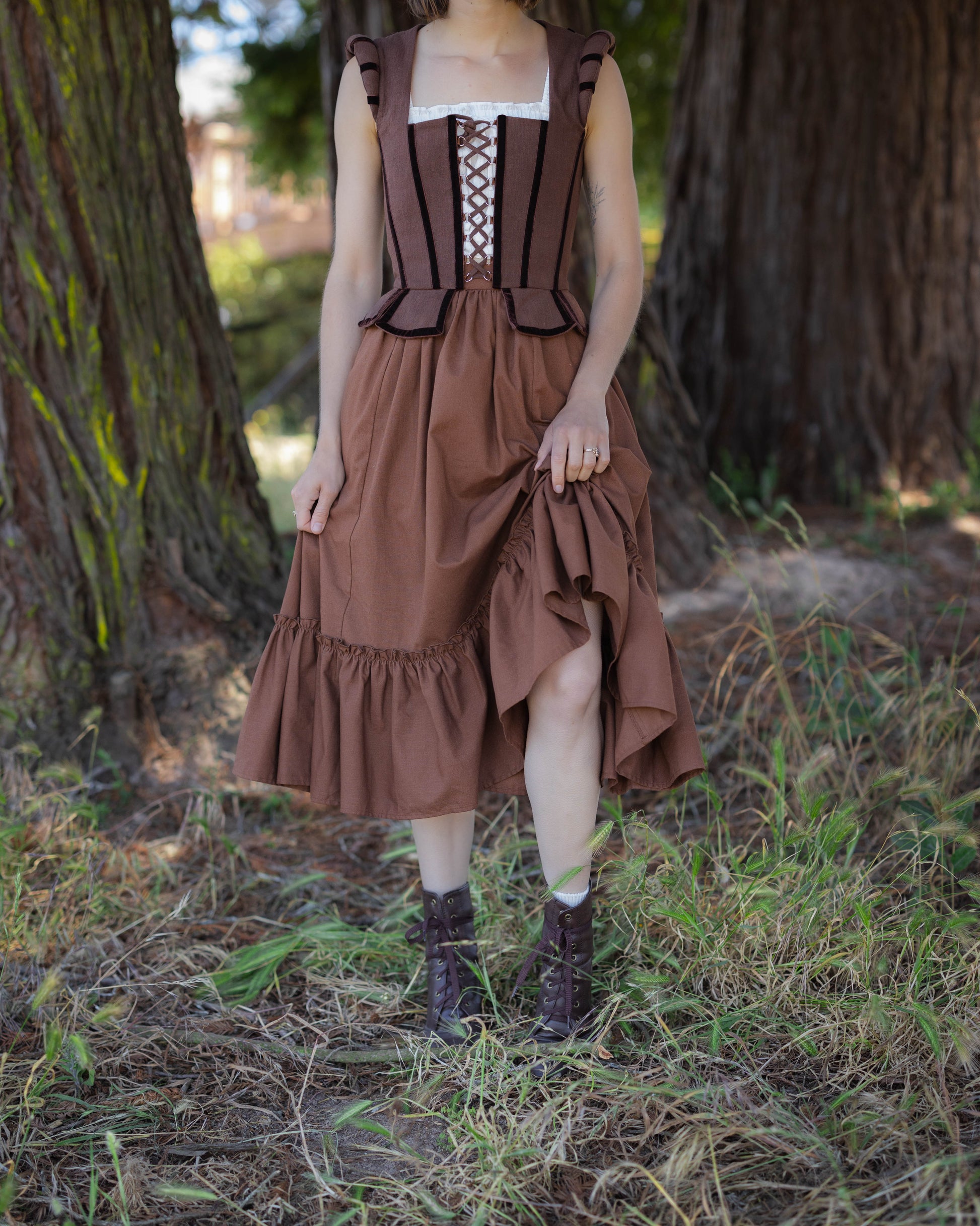 Woman wearing a brown linen ruffle skirt and corset standing outdoors on grass near large tree trunks
