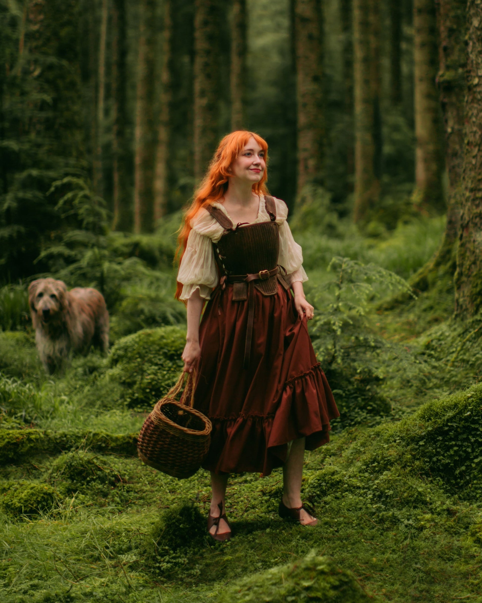Woman with red hair wearing rust linen midi skirt and cream blouse standing in green forest with wicker basket and dog nearby