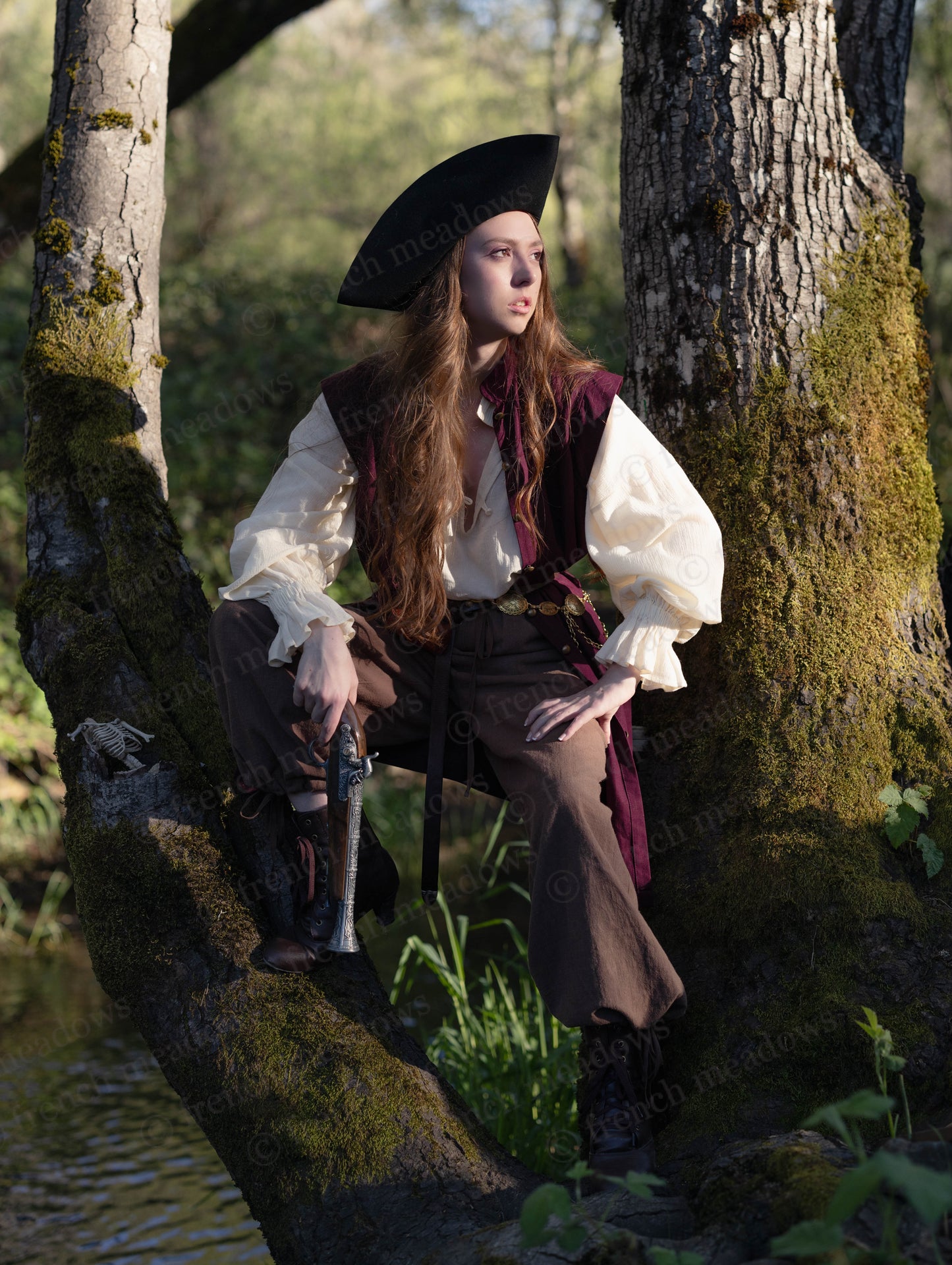 Young person wearing Burgundy Pirate Vest over a white shirt and dark pants, seated on a mossy tree by water.