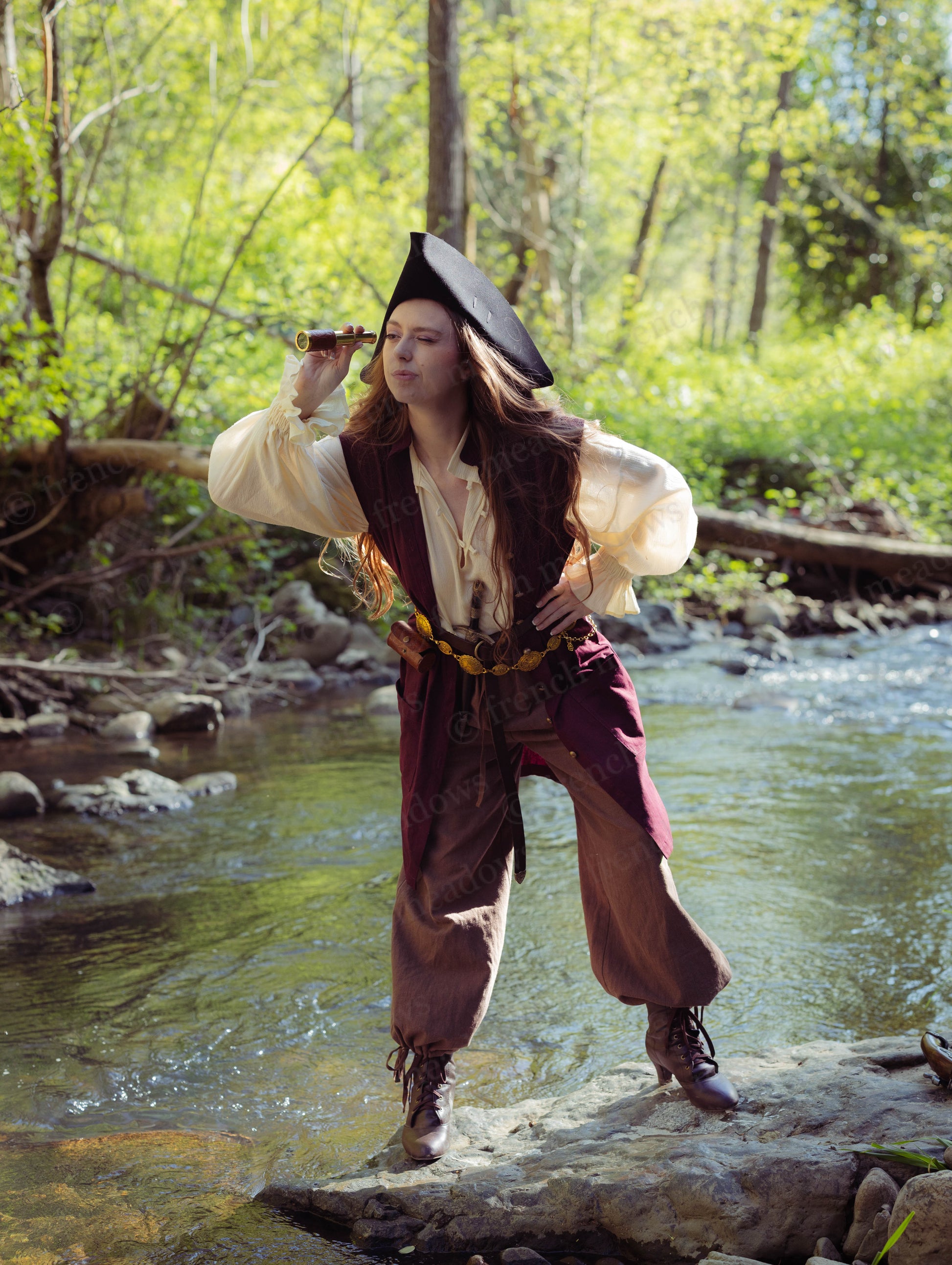 Person wearing rustic pirate pants with a vest and hat, standing by a forest stream looking through a spyglass