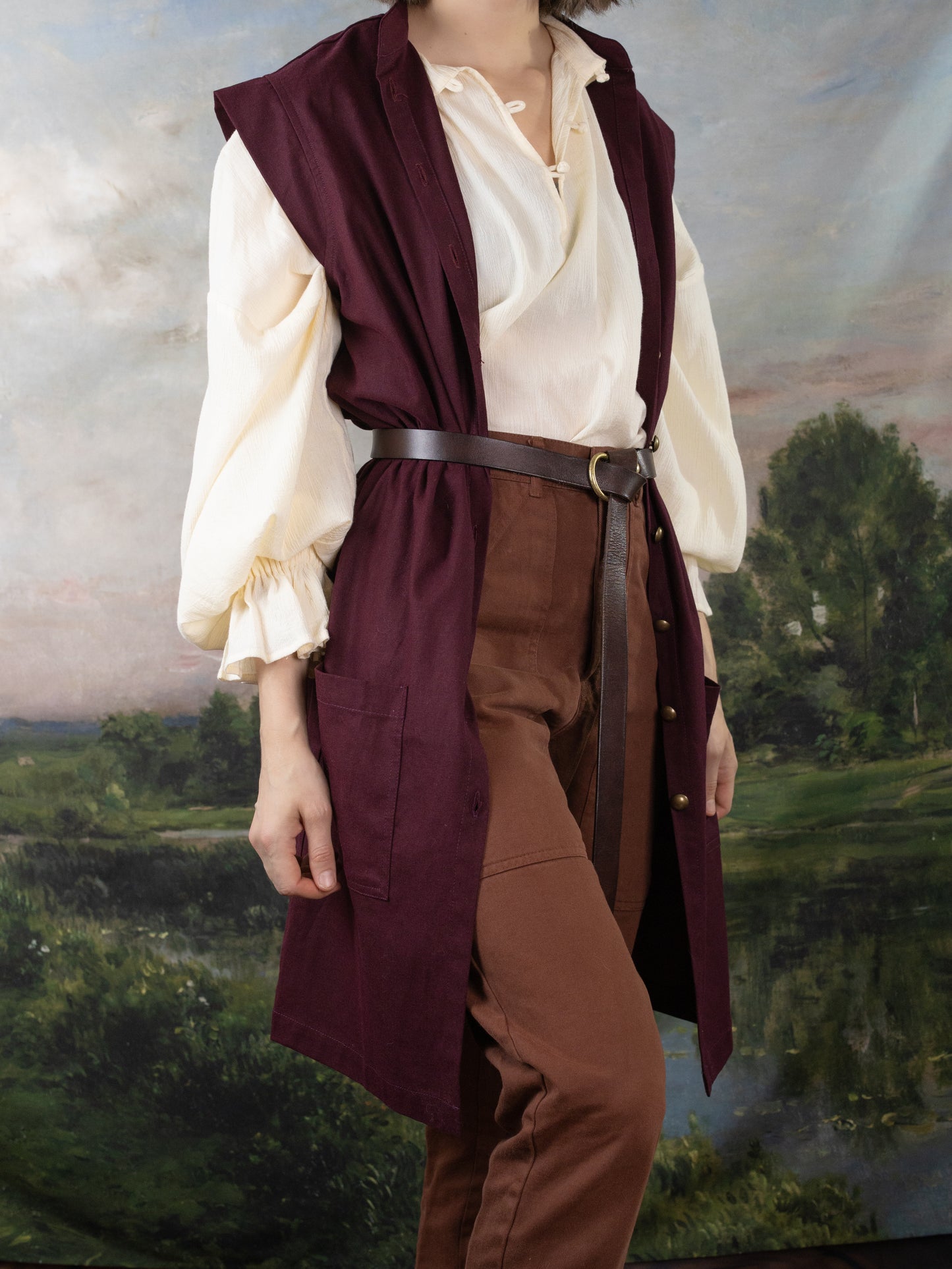 Burgundy Pirate Vest layered over a cream Darcy Shirt with a leather waist belt in a renaissance fair setting.