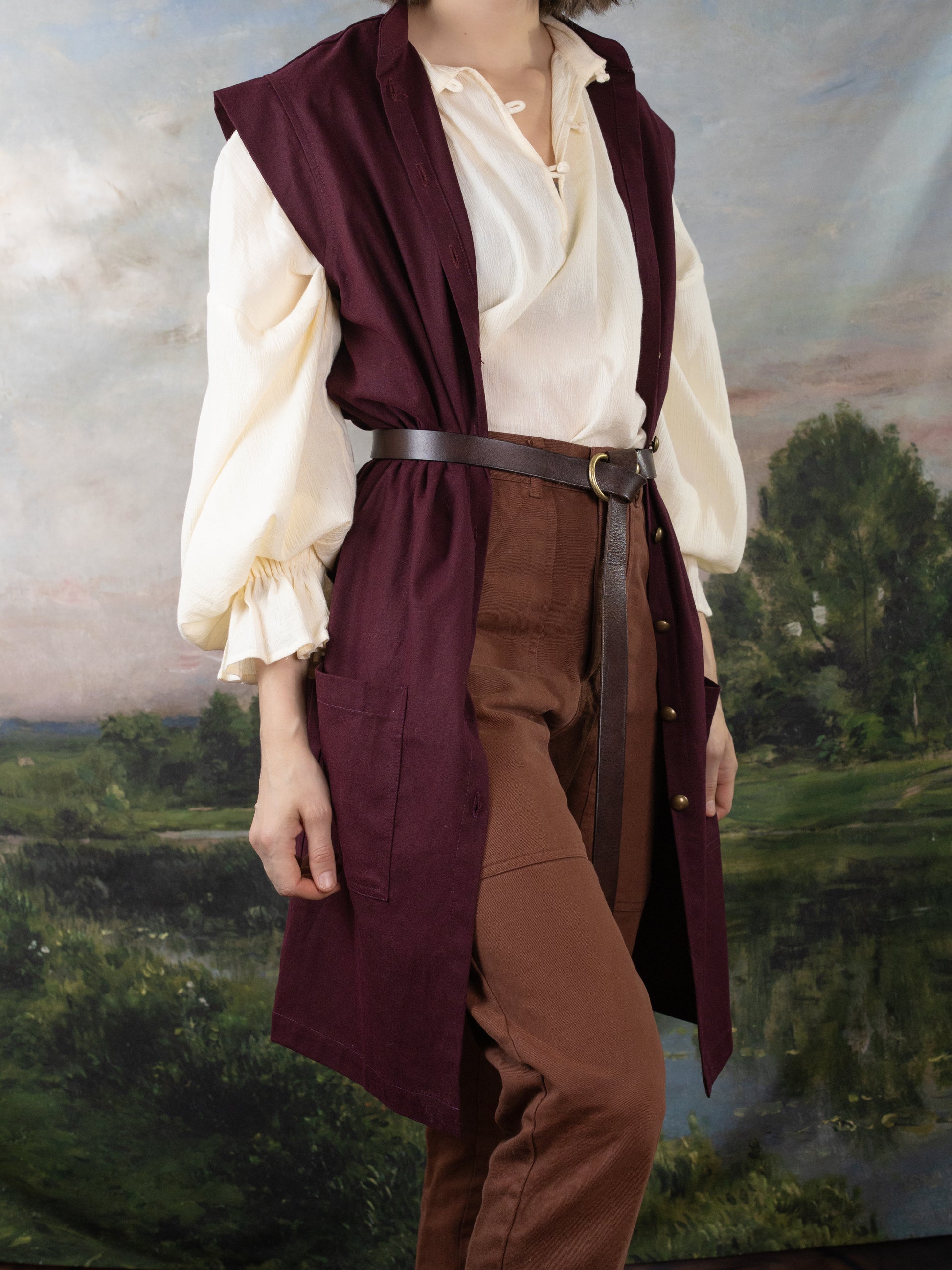 Burgundy Pirate Vest layered over a cream Darcy Shirt with a leather waist belt in a renaissance fair setting.