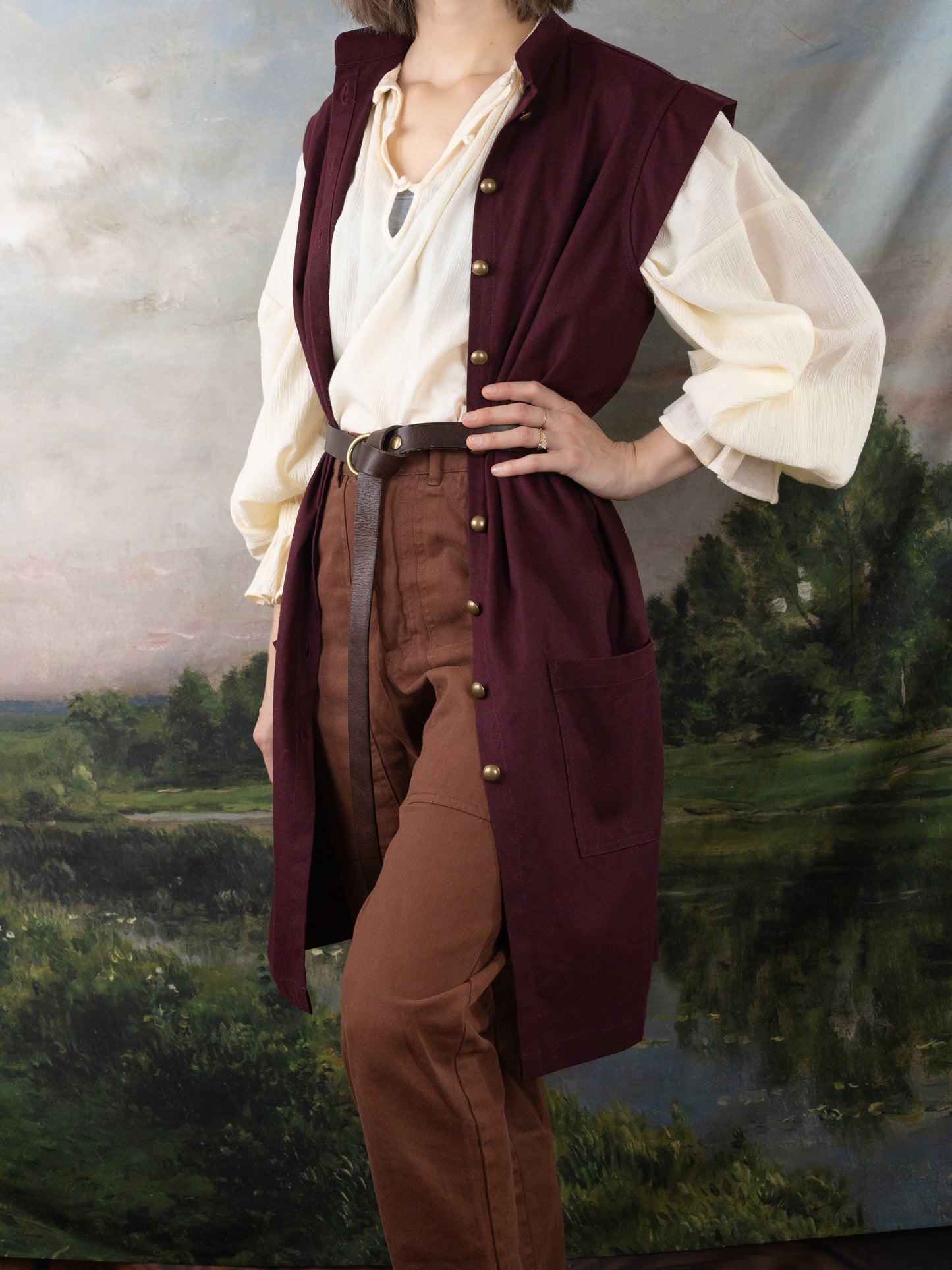 Woman wearing a Burgundy Pirate Vest over a cream shirt and brown pants with a leather belt standing before a painted backdrop.