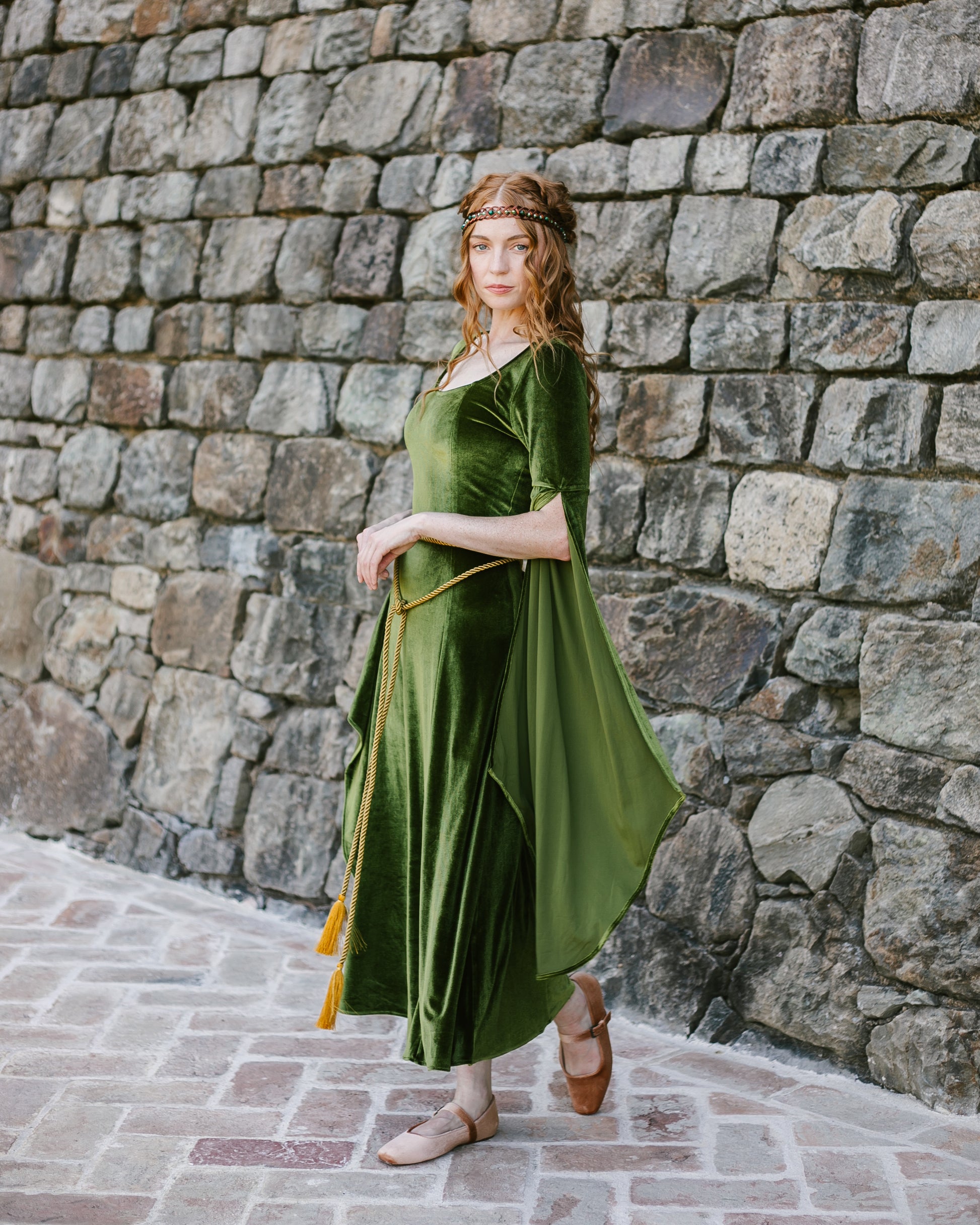 Woman wearing a chartreuse velvet medieval dress with bell sleeves and back lacing standing against a stone wall.