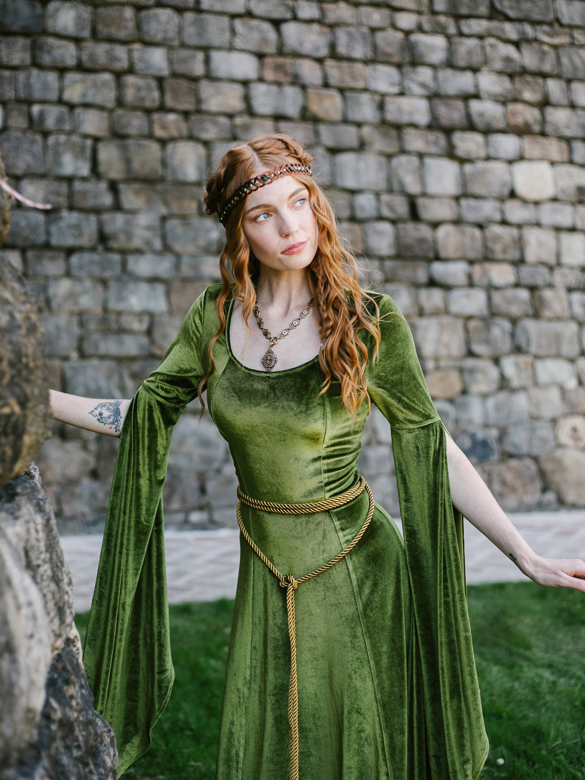 Woman in a chartreuse velvet medieval bell sleeve dress with braided belt and headpiece against stone wall background