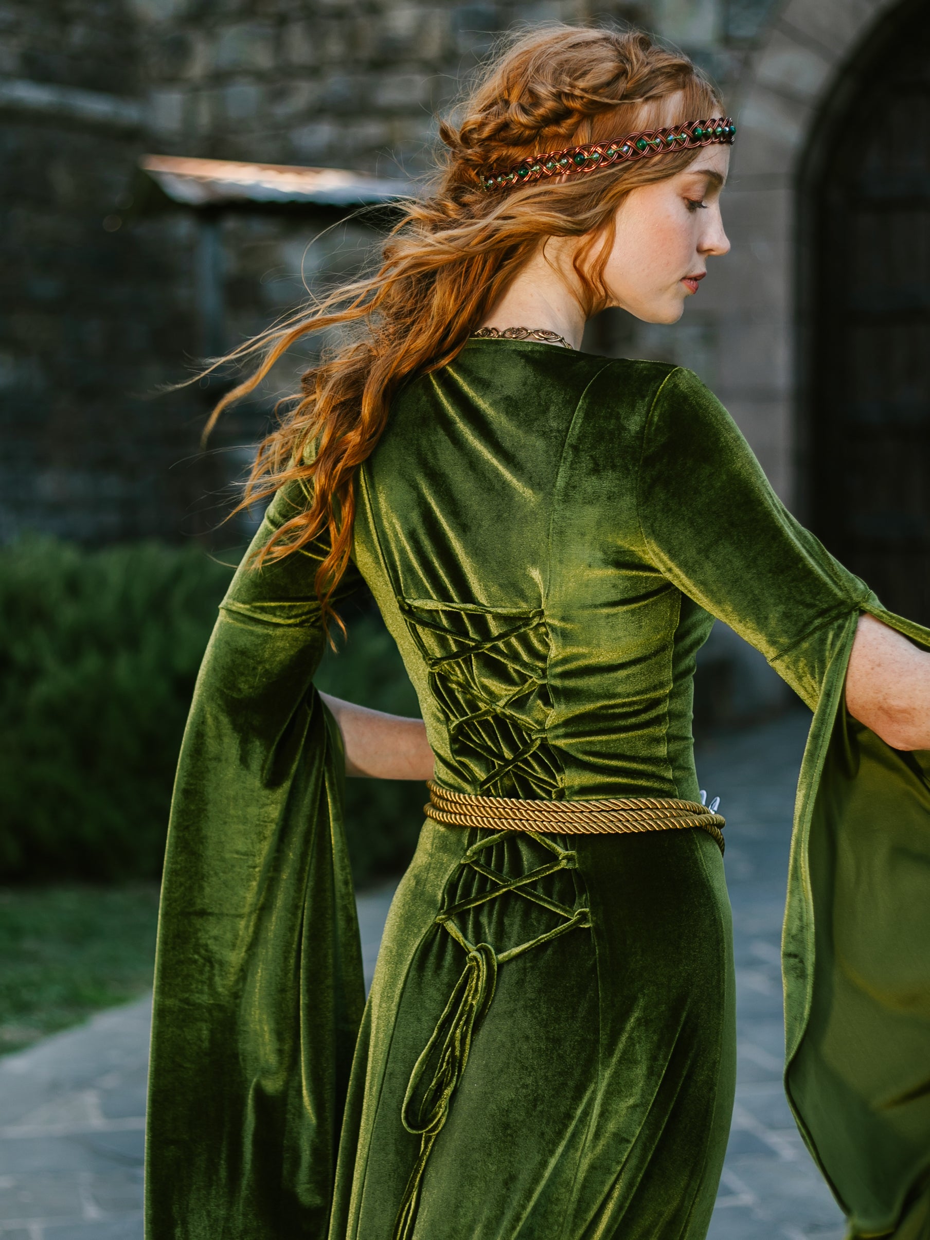 Woman wearing a chartreuse velvet medieval dress with bell sleeves and adjustable back lacing, standing outdoors by stone walls