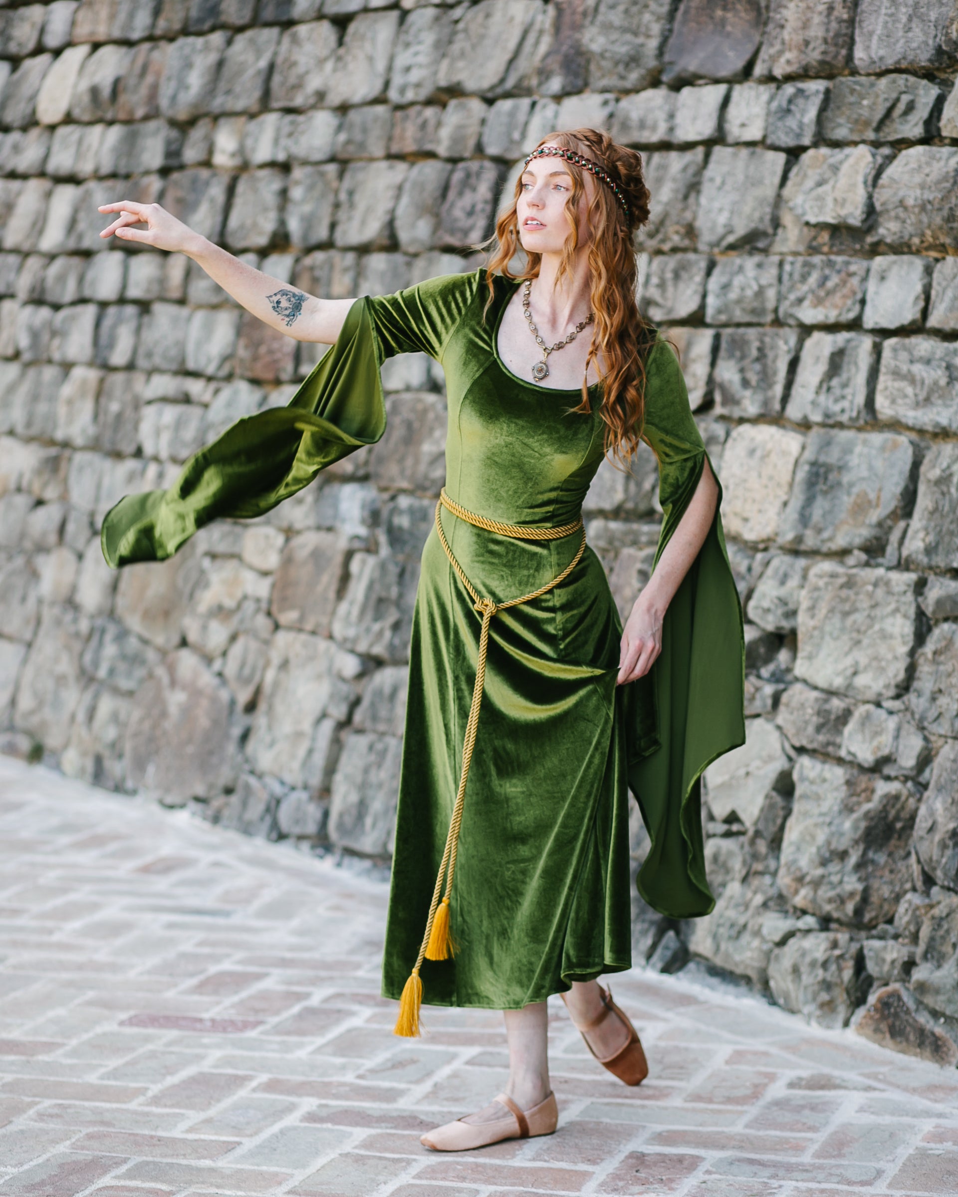 Woman in green medieval gown wearing a long golden rope belt with tassel ends, standing by a stone wall