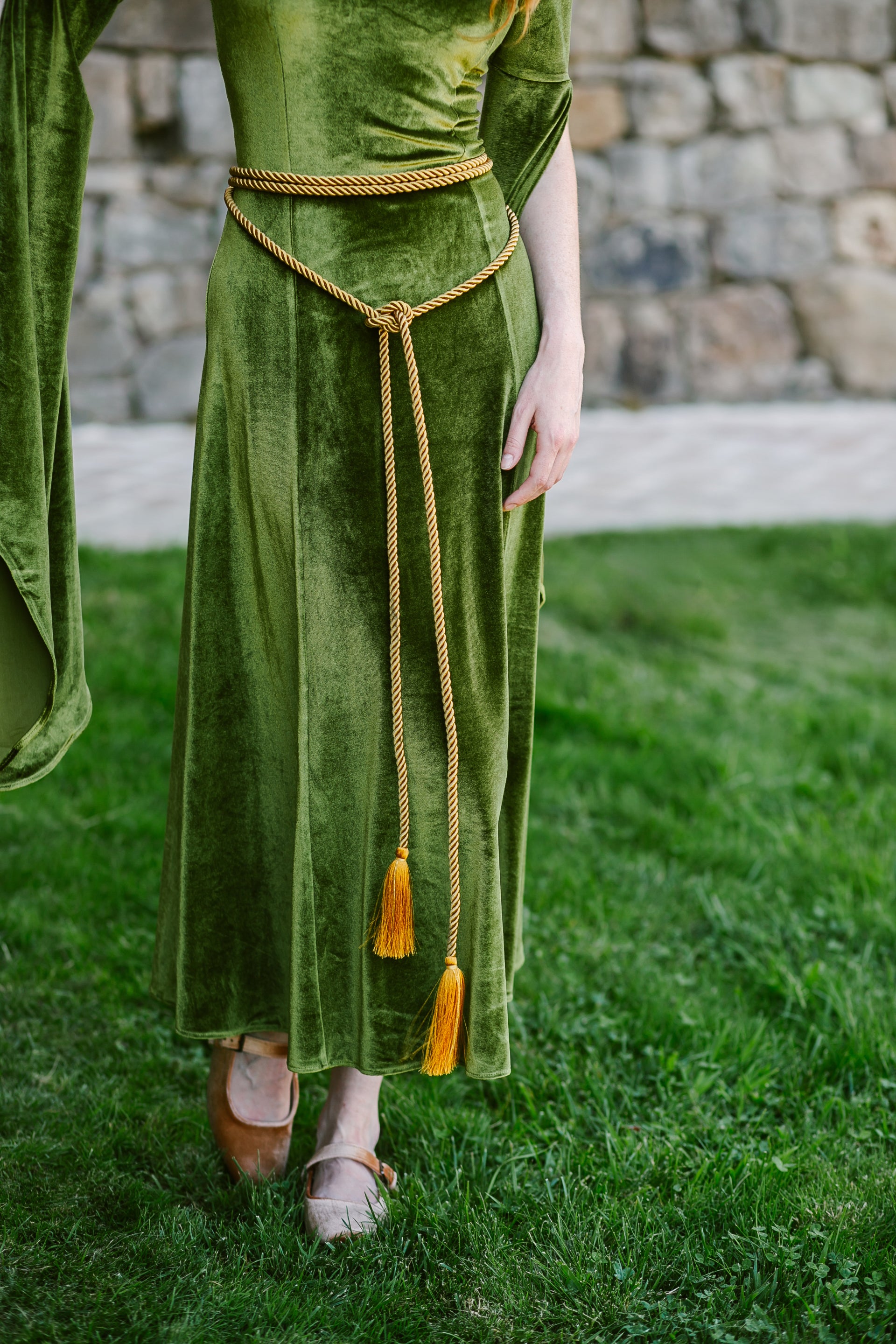Golden medieval rope belt with tassel ends wrapped around a green velvet gown waist outdoors near stone wall