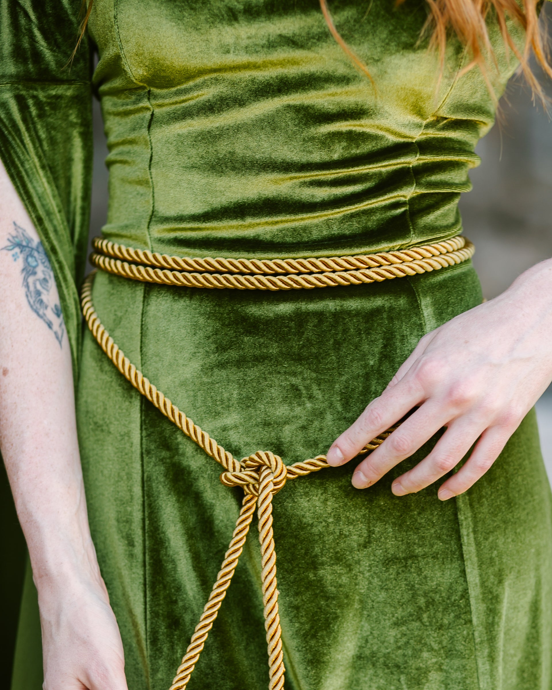 Close-up of a golden medieval rope belt tied around the waist over a green velvet dress with tassel ends visible.