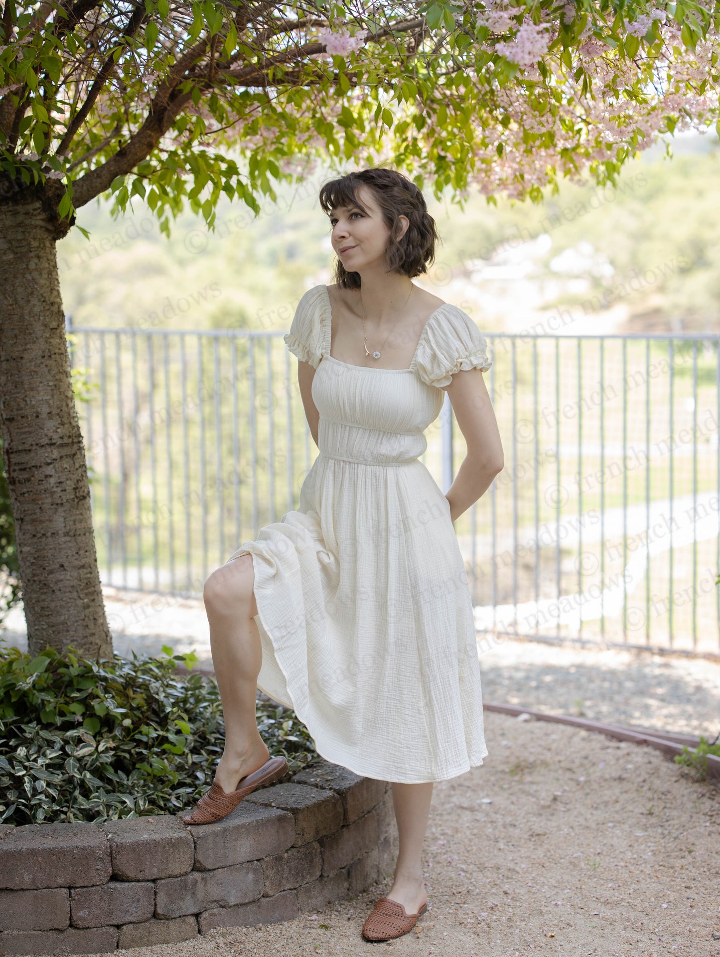 Ivory Cotton Milkmaid Dress with square neckline and puff sleeves shown in garden setting under flowering tree branches