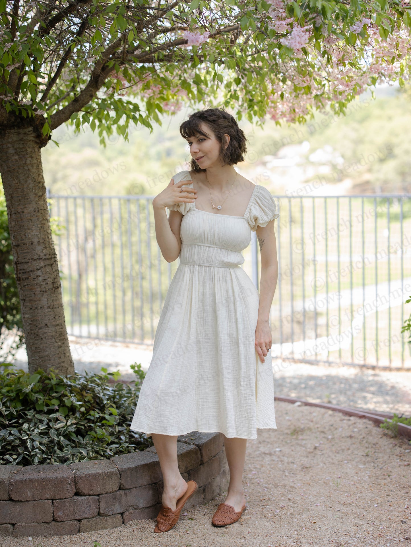 Ivory Cotton Milkmaid Dress styled under blooming tree with puff sleeves and square neckline, shown in outdoor garden setting