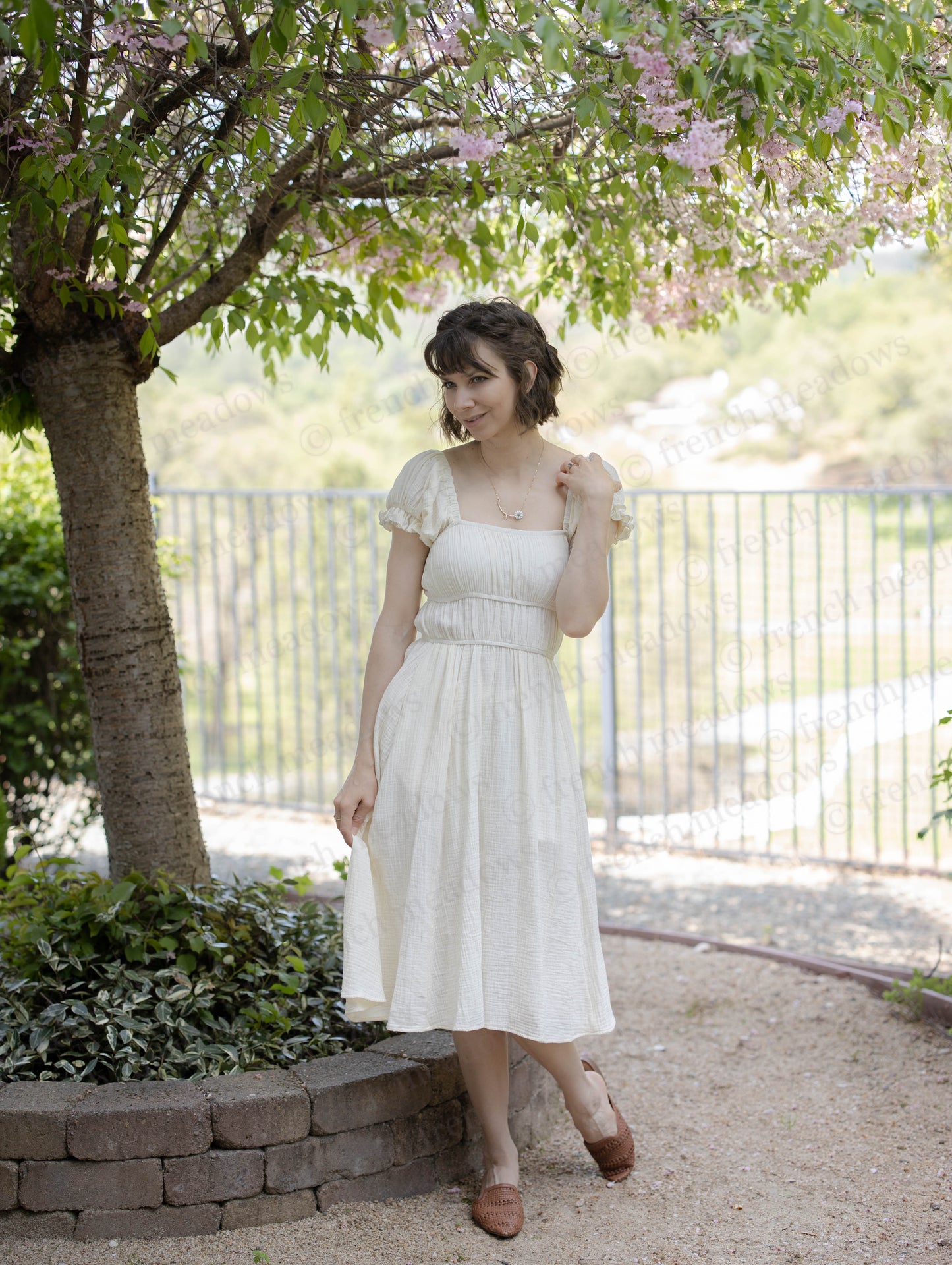 Ivory Cotton Milkmaid Dress with square neckline and ruched bodice shown outdoors under blooming tree with garden backdrop