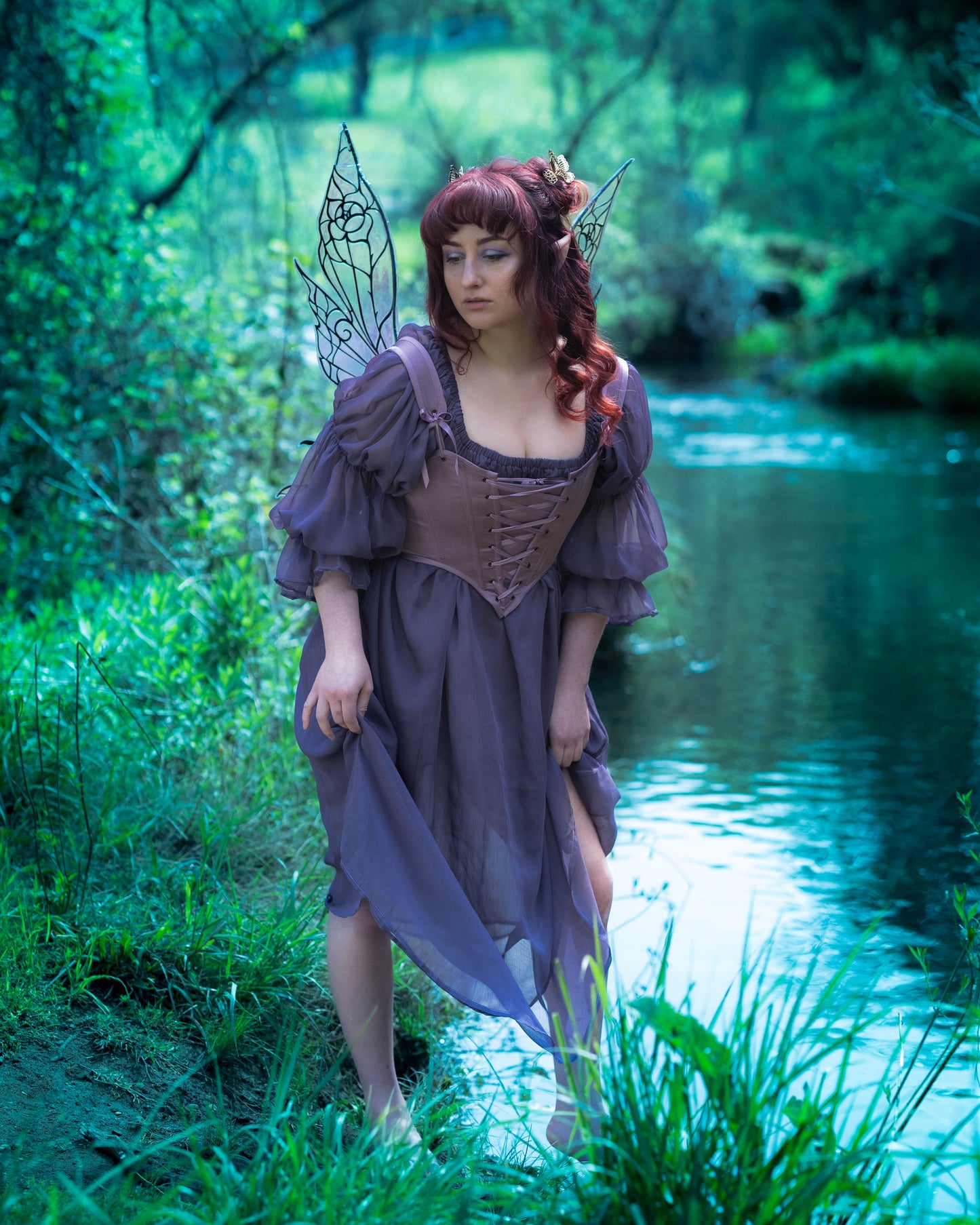 Woman wearing a Purple Dusk Satin Corset Top with lacing and fairy wings standing by a forest stream.