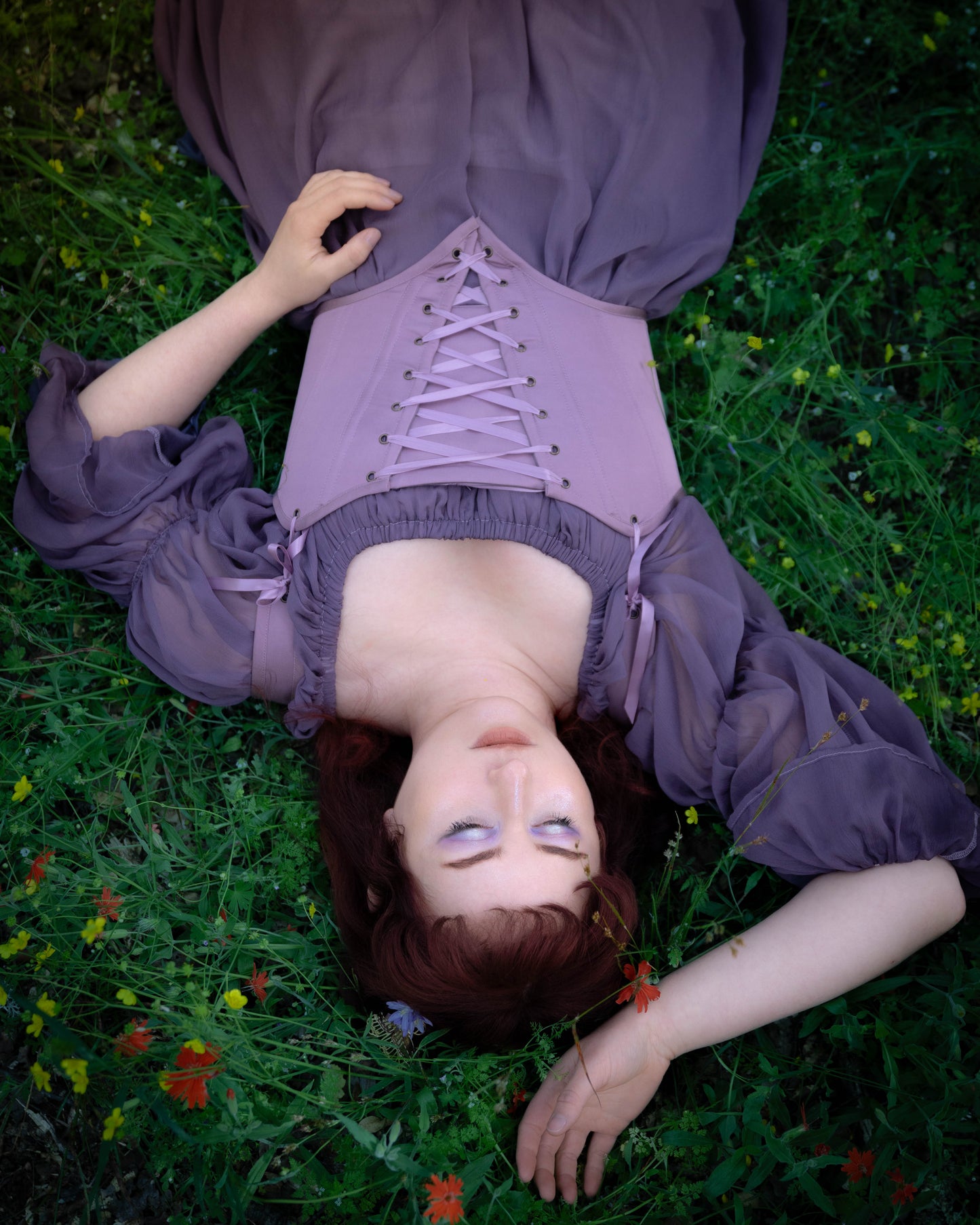 Woman lying in grass wearing Purple Dusk Satin Corset Top with front lacing and sheer sleeves in dusky purple hue.