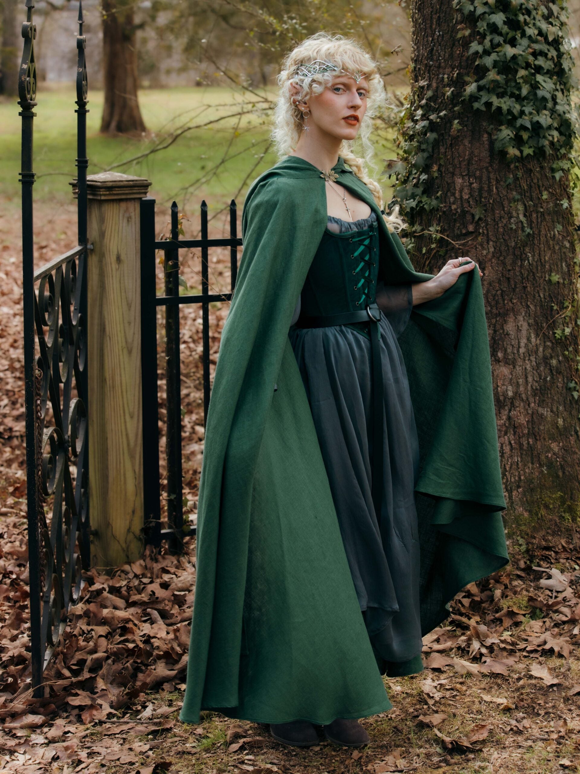 Elegant forest green Linen Renaissance Cloak shown in autumn garden setting with wrought iron fence and fallen leaves