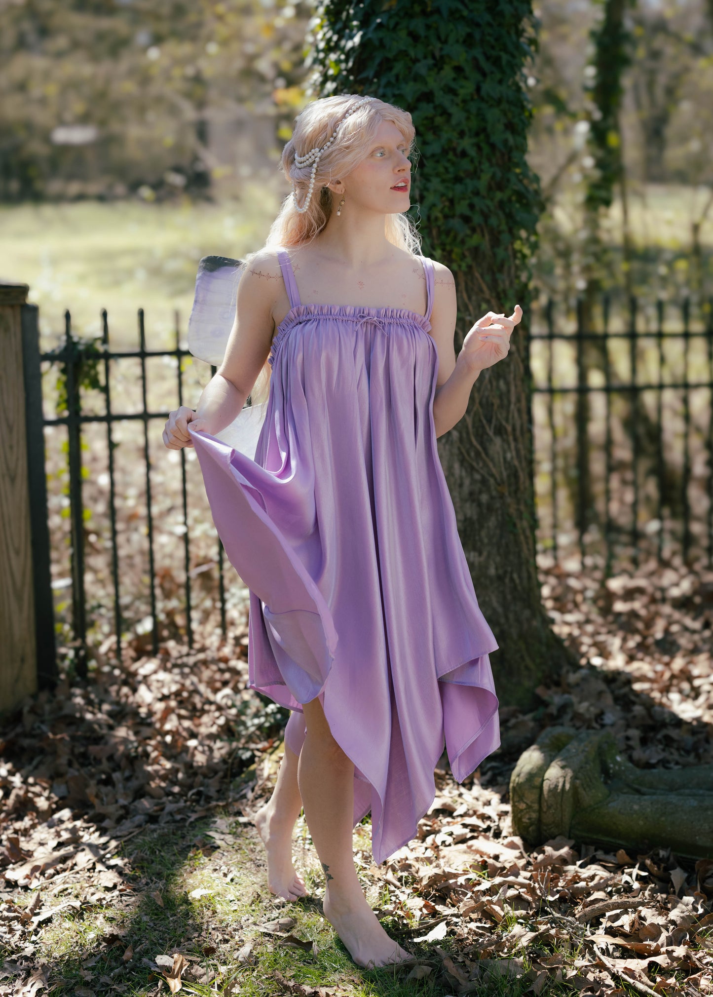 Woman wearing a flowing lavender Flutter Fairy Dress with handkerchief hem standing barefoot outdoors near a tree.