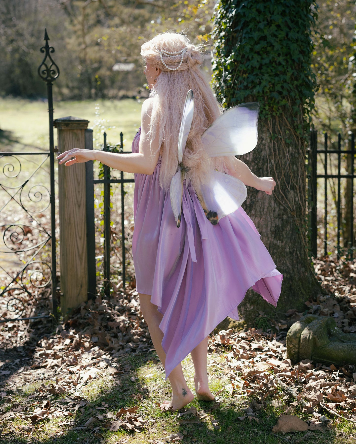 Woman wearing a lilac Flutter Fairy Dress with handkerchief hem and fairy wings outdoors by a tree and gate.