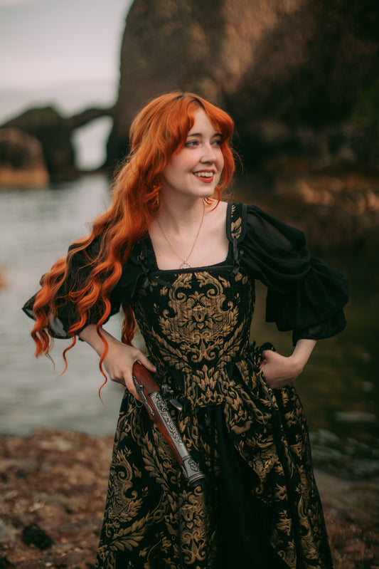 Young woman with long red hair wearing black and gold chenille Tudor stays corset holding an ornate vintage pistol outdoors