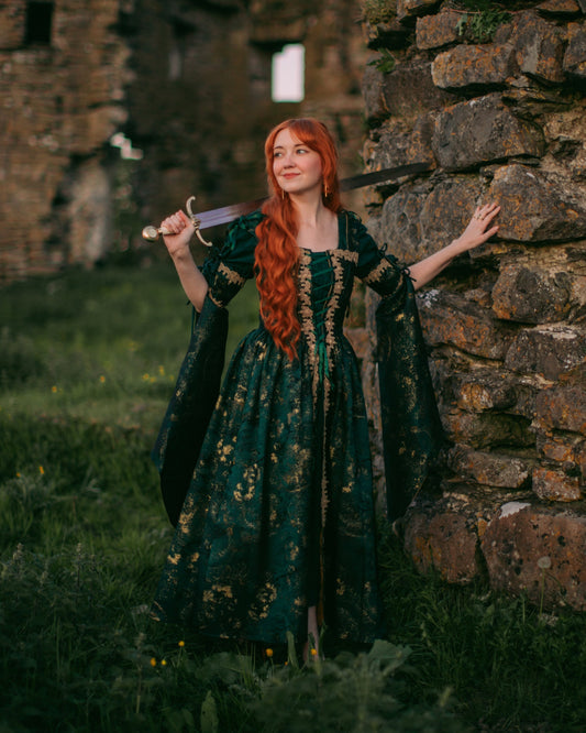 Woman in medieval ruins wearing a Dark Green Jacquard Corset Dress with gold accents and removable bell sleeves.