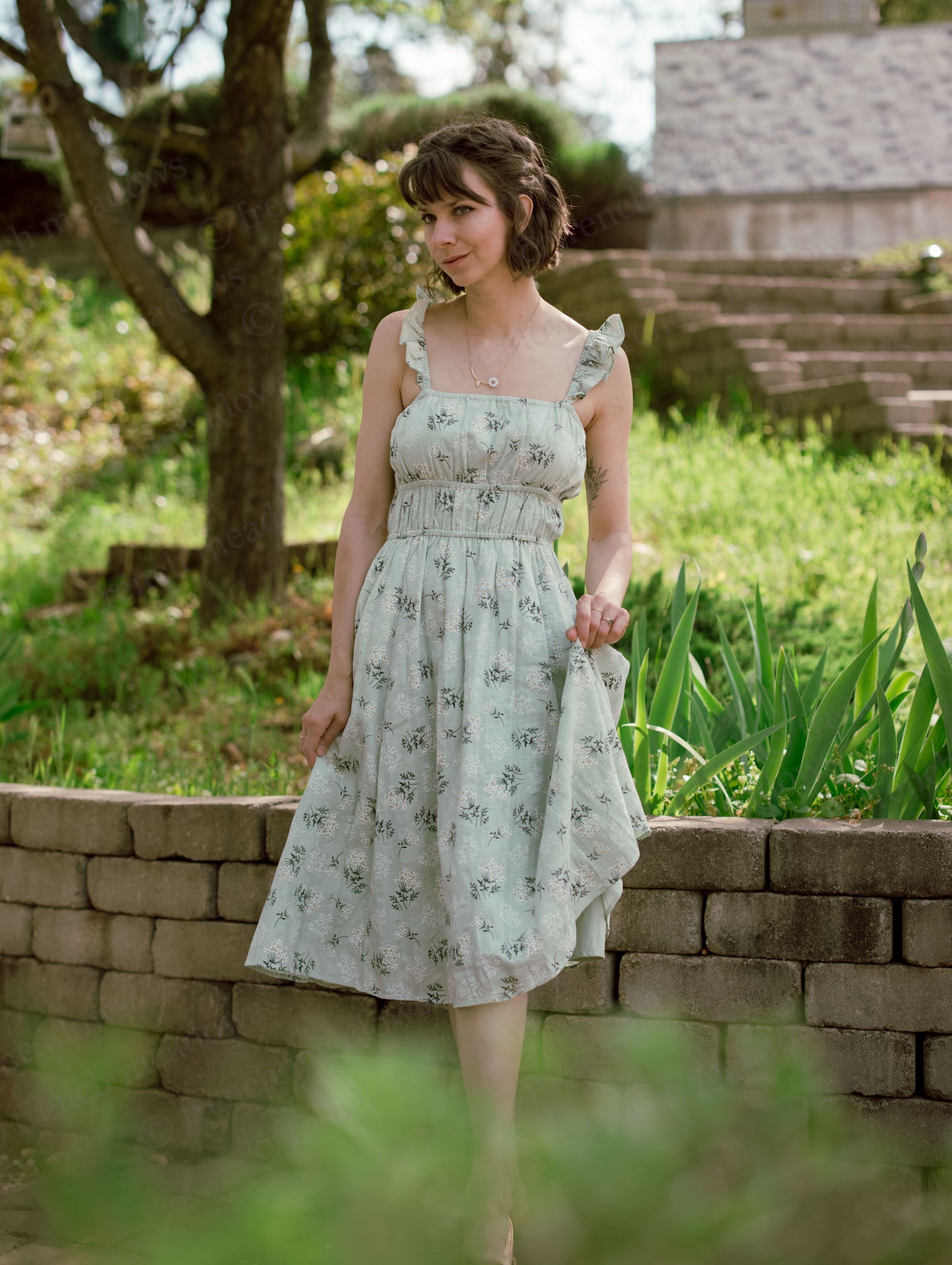 Woman wearing a Floral Milkmaid Dress with ruched bodice and A-line skirt standing outdoors by a stone wall.